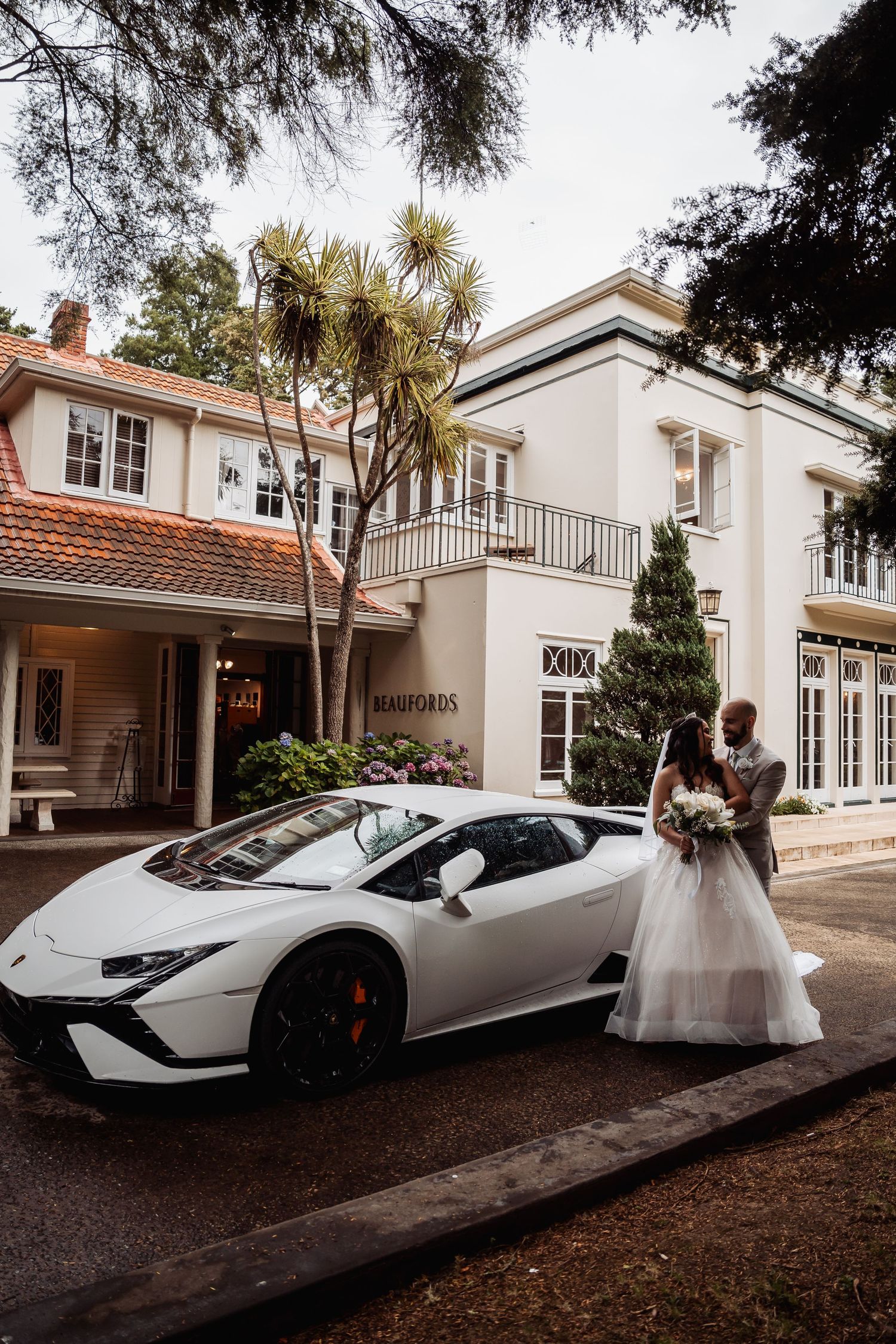 Wedding couple poses beside white Lamborghini in front of elegant manor venue entrance.