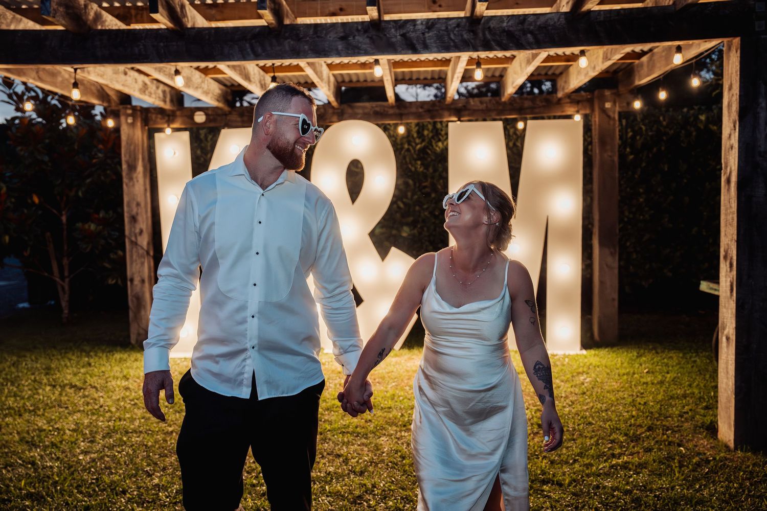Newlyweds hold hands in front of illuminated marquee letter signs V&M during their evening outdoor wedding.
