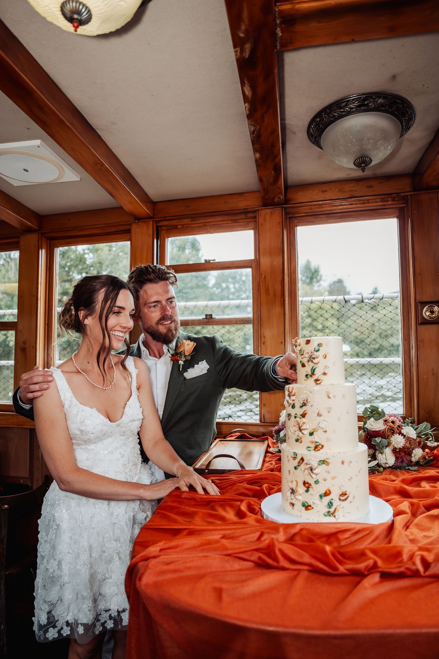 Bride and groom cut elegant floral wedding cake on boat decorated with orange satin tablecloth.