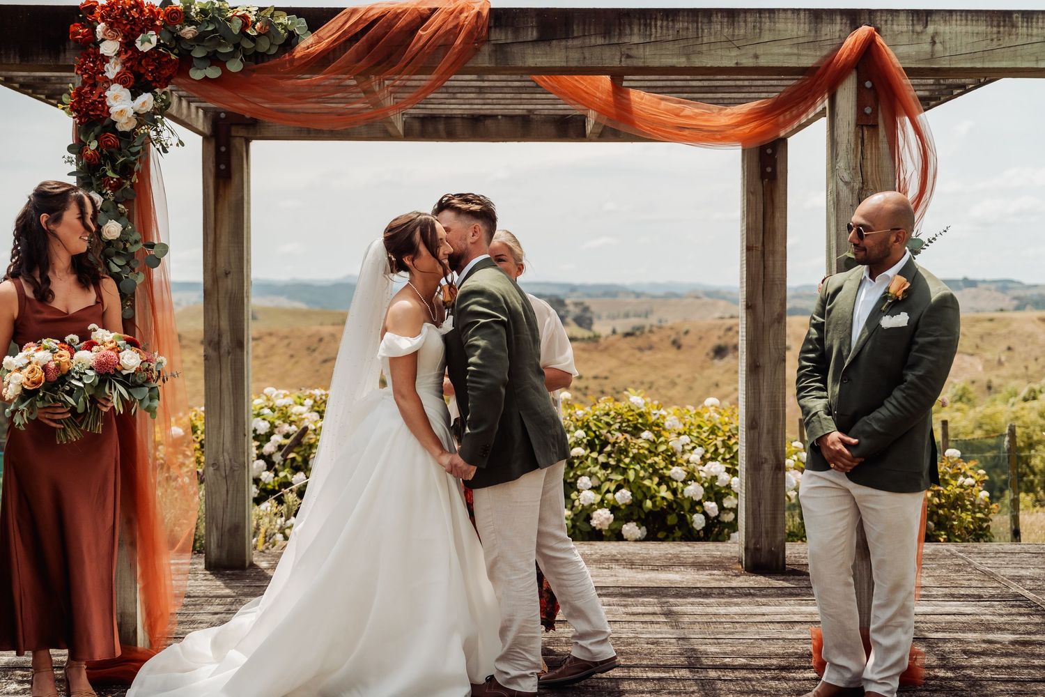 Bride and groom sharing first kiss at outdoor ceremony under rustic wooden arch with orange draping.