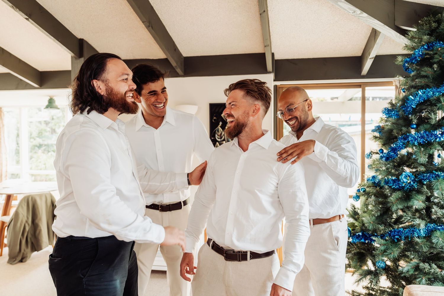 Four men in white shirts laughing and celebrating together near a decorated Christmas tree at a wedding event.