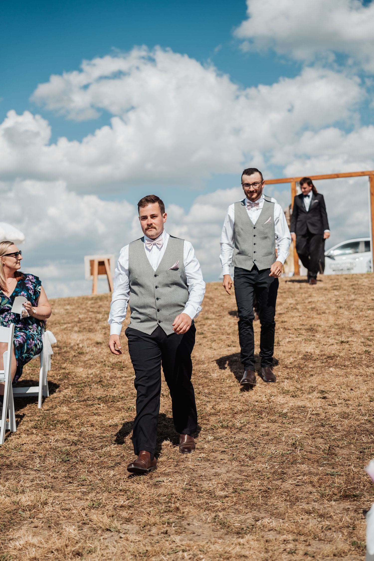 Two groomsmen in grey vests and black trousers walk down outdoor wedding aisle under a cloudy sky.