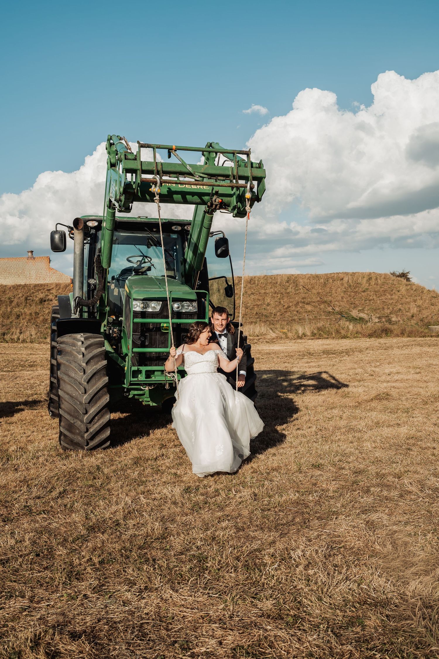 Bride and groom pose together in front of a green John Deere tractor in a golden harvested field under blue sky.