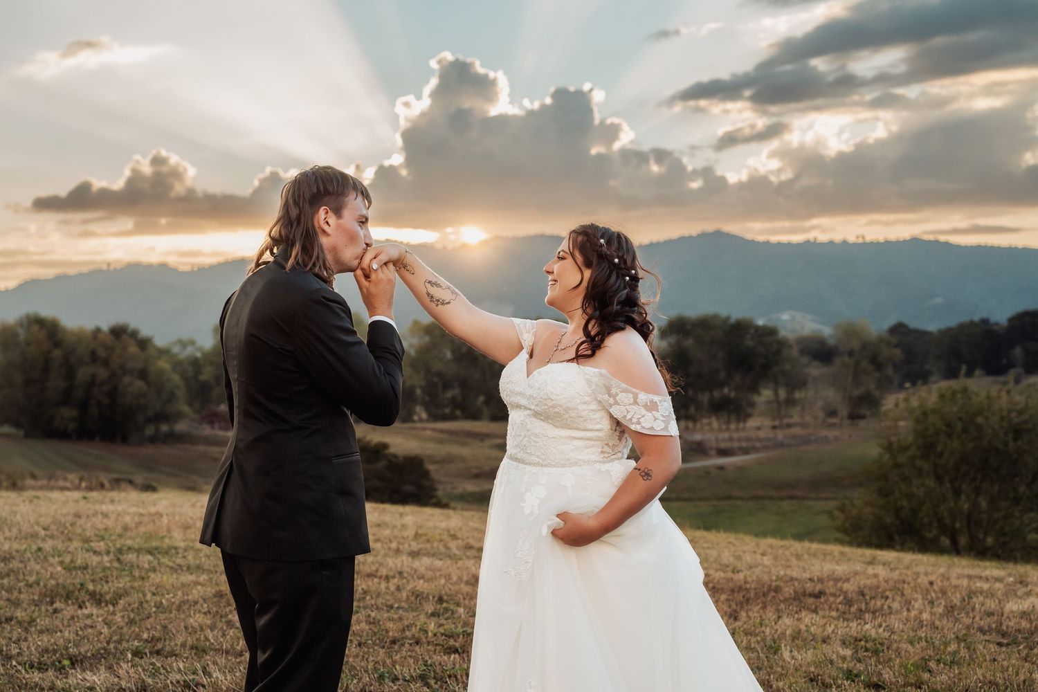Groom kisses bride's hand at golden sunset over mountain meadow with dramatic rays breaking through clouds.