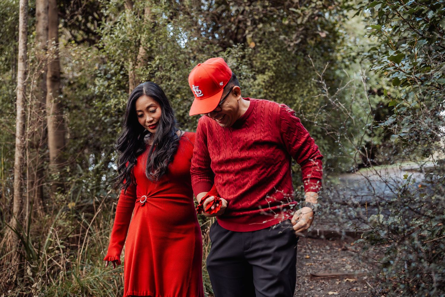 Couple in matching red outfits walking hand in hand through a lush green forest path.
