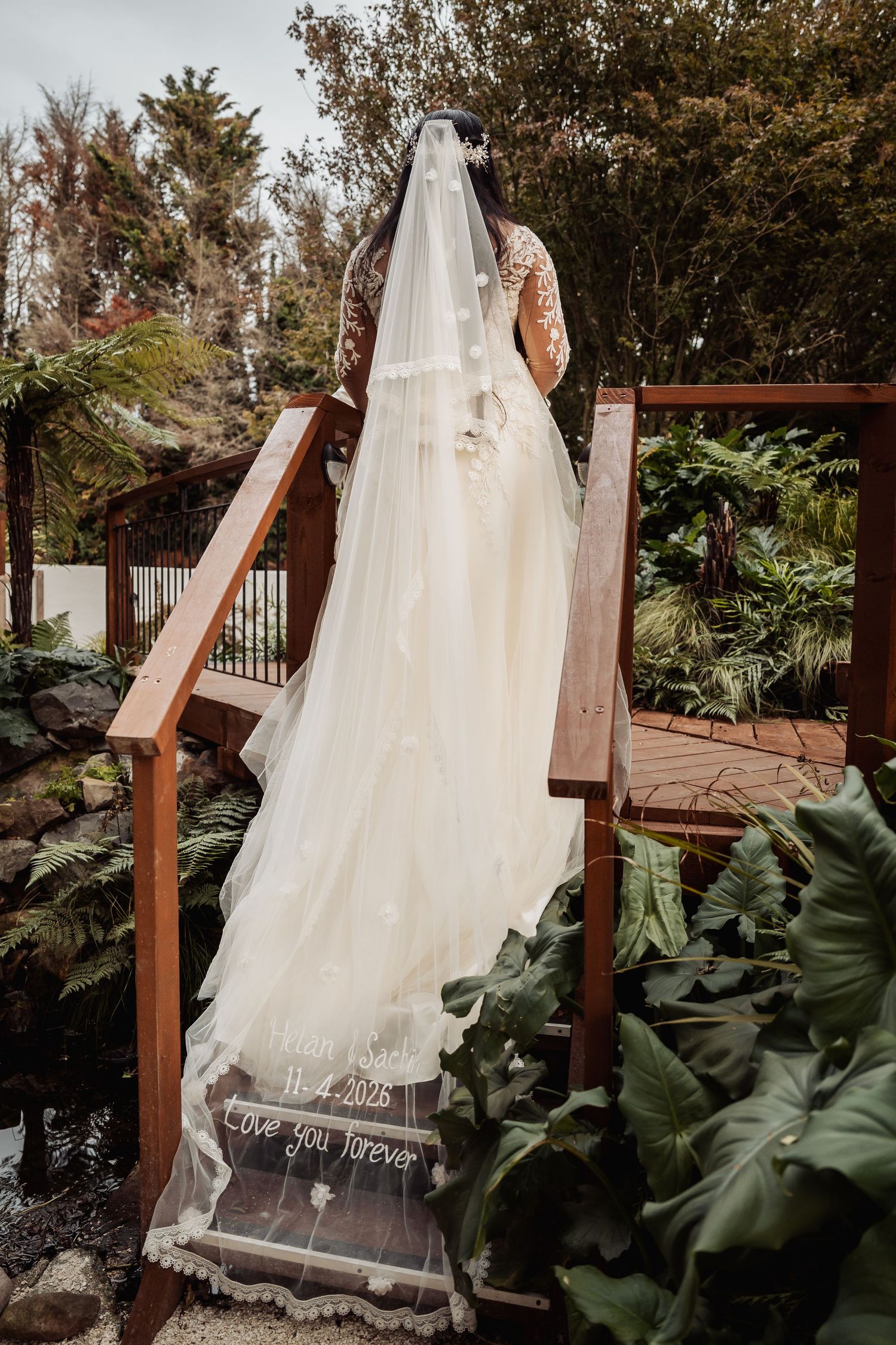 Bride in lace wedding dress with personalized veil reading Love You Forever on wooden garden bridge.