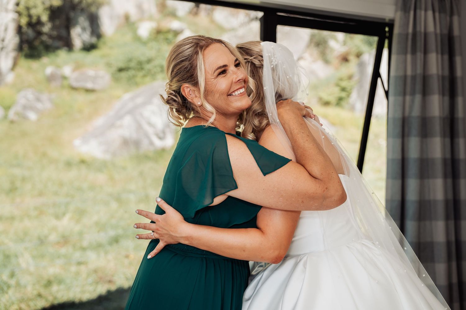Bride in white gown and veil embracing smiling bridesmaid in green dress outdoors.