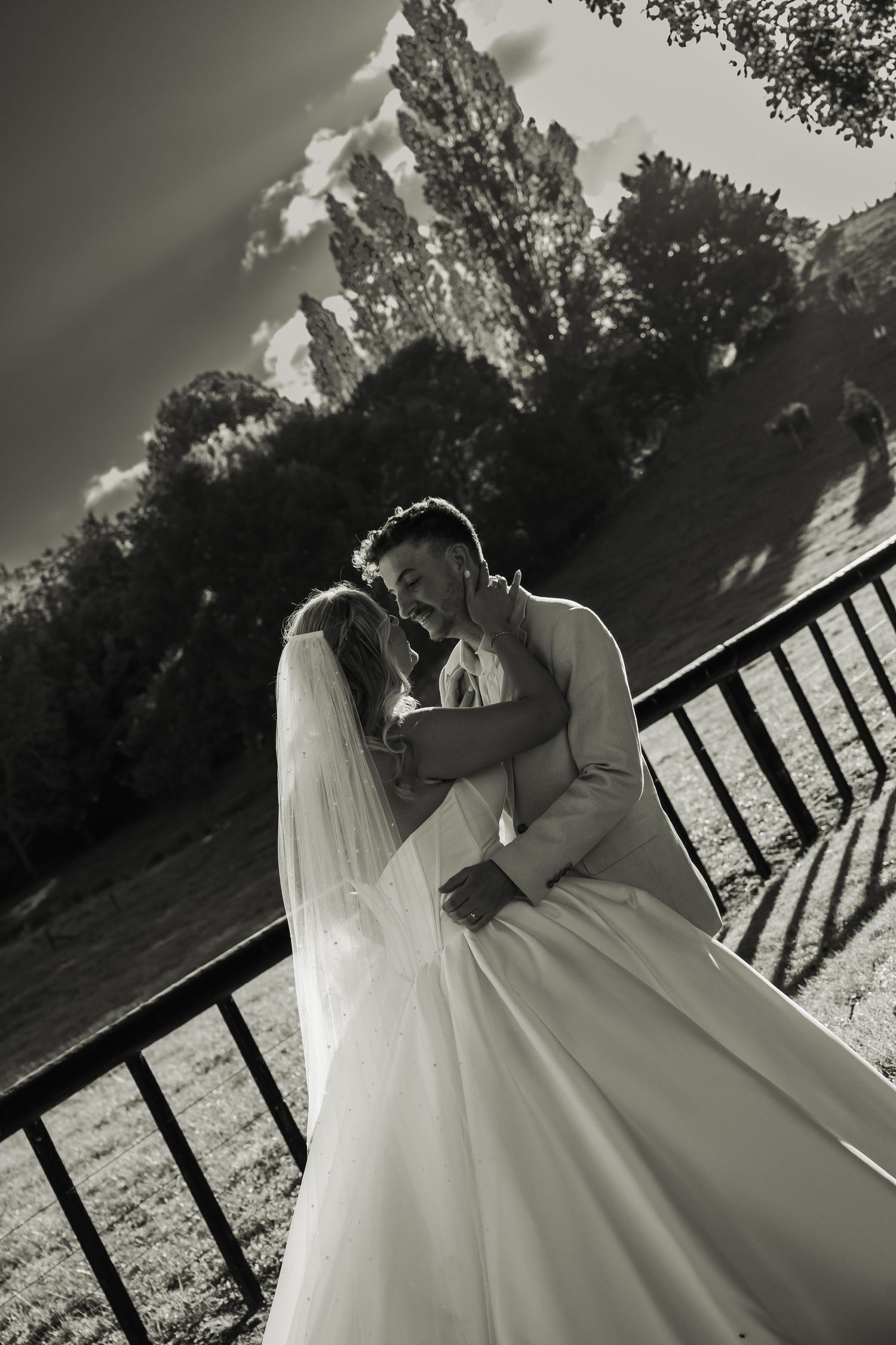 Black and white romantic portrait of bride and groom embracing near fence in outdoor setting.