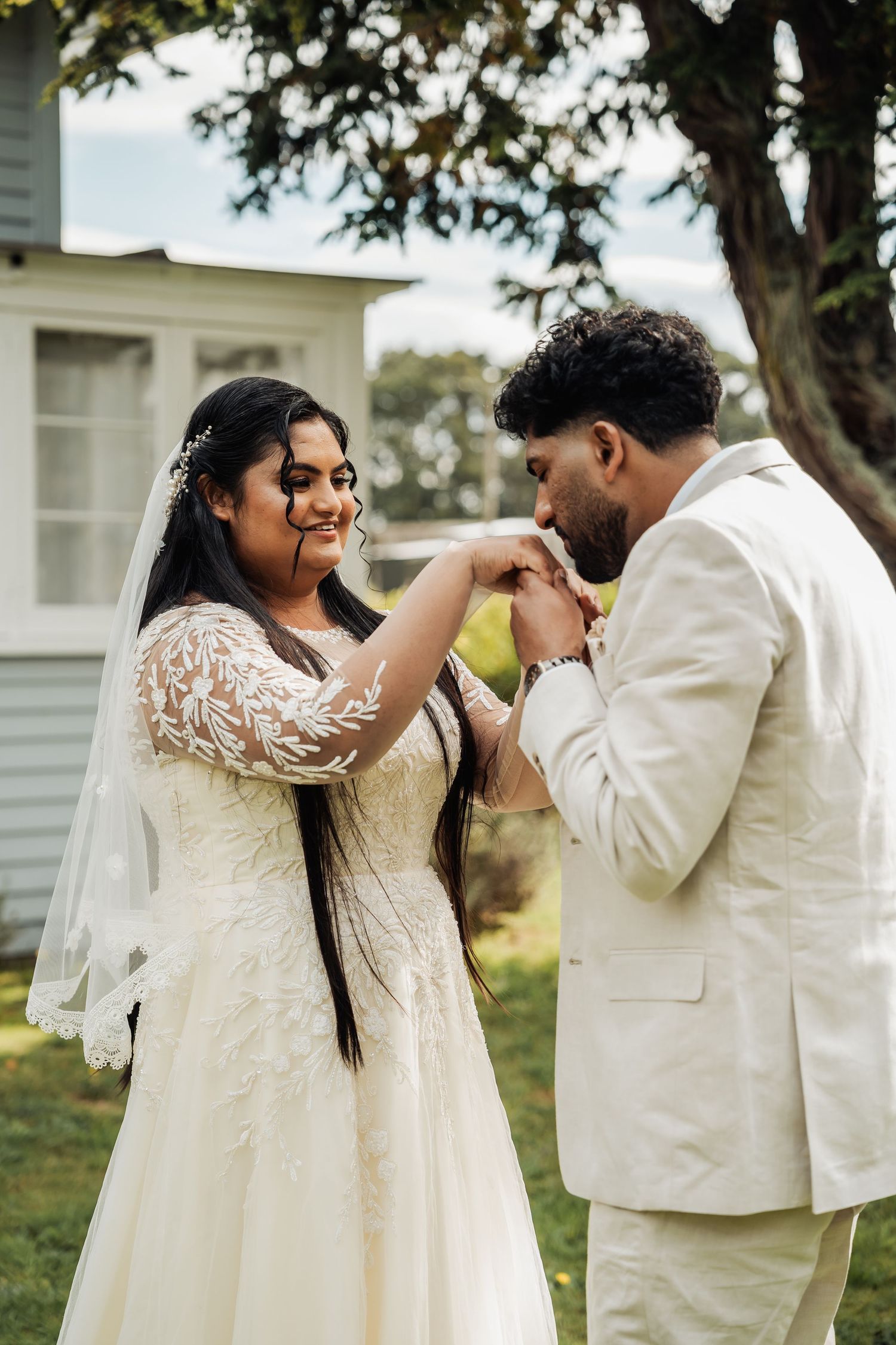 Groom in white suit kissing bride's hand outdoors during wedding ceremony.