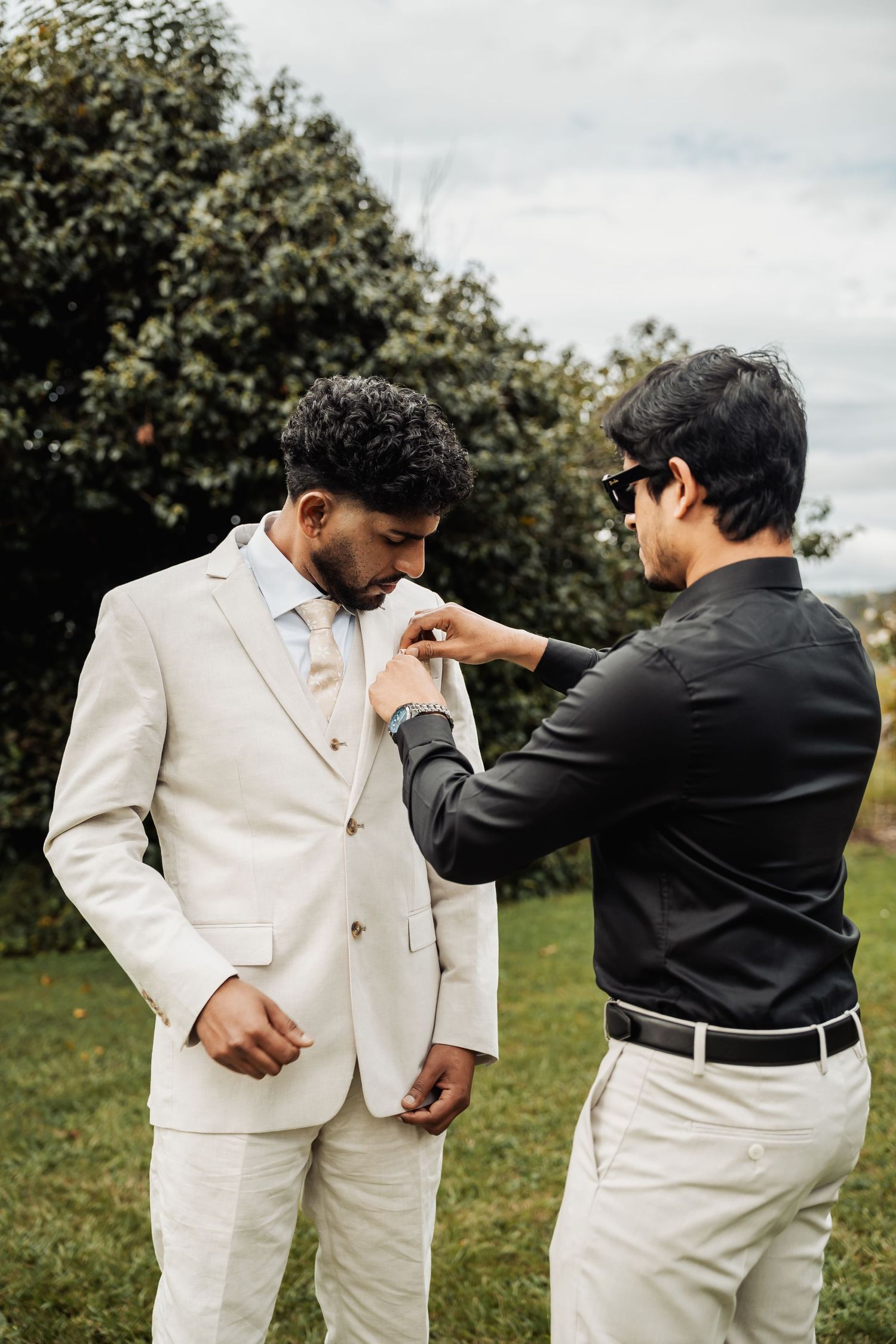 A man in a black shirt pins a boutonniere on a groom wearing a cream suit outdoors.