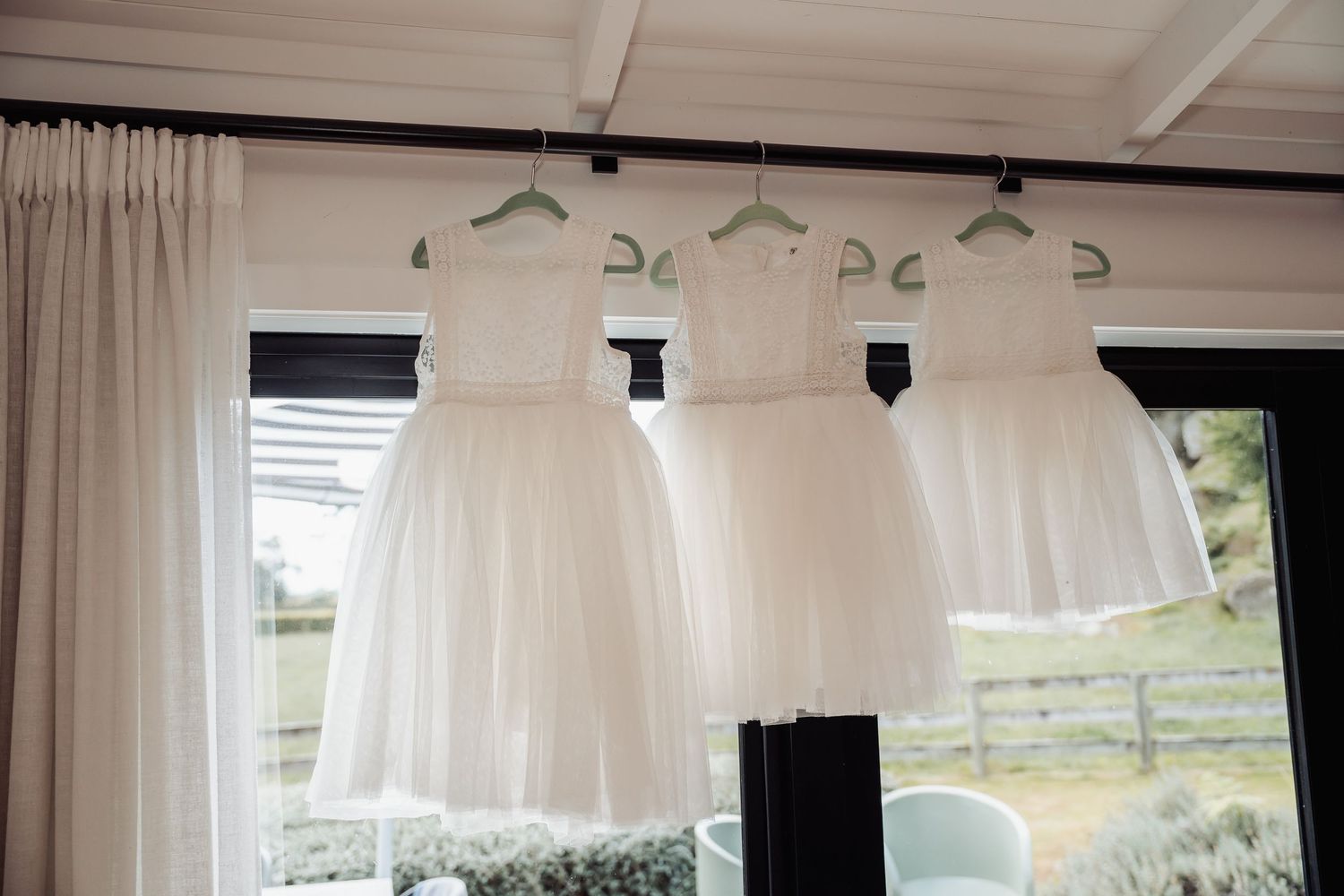 Three white flower girl dresses hanging on green hangers in front of a sunlit window.