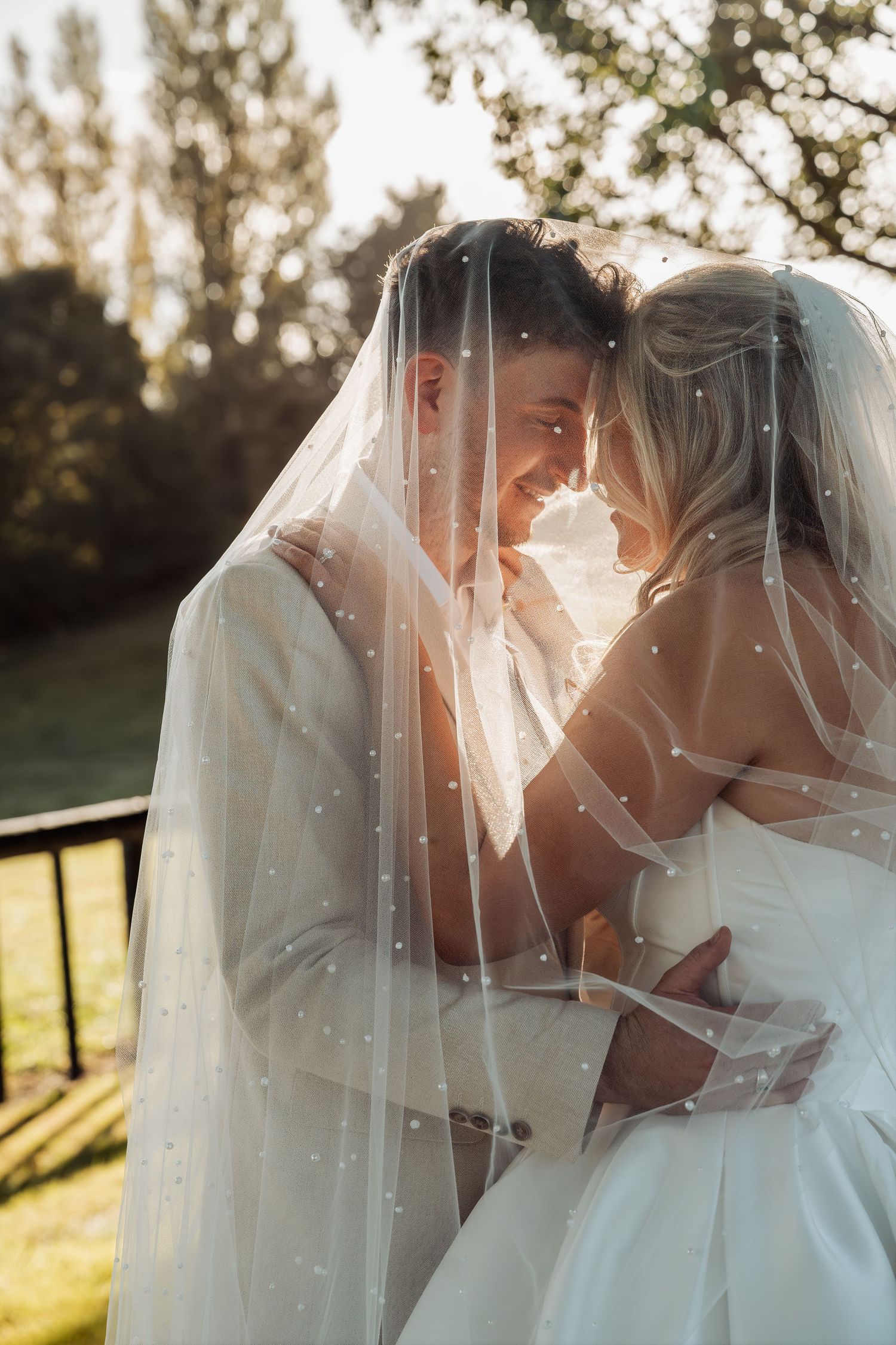 A bride and groom share an intimate moment together under a pearl-dotted veil in soft golden sunlight.