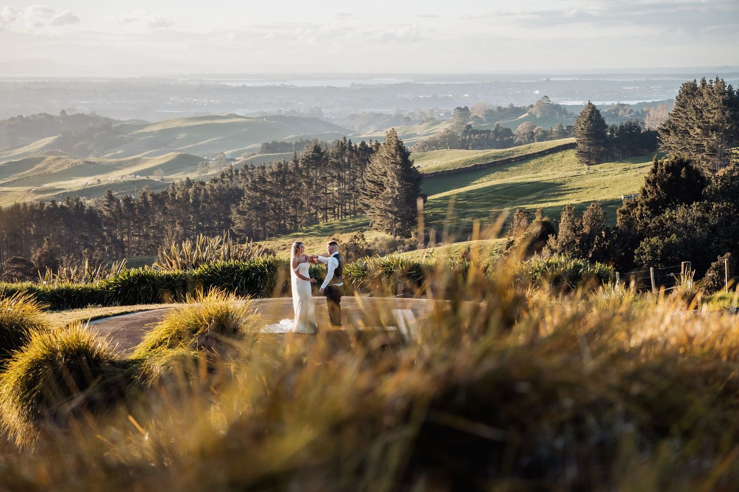 Wedding couple standing on scenic hilltop road surrounded by rolling green New Zealand countryside landscape.