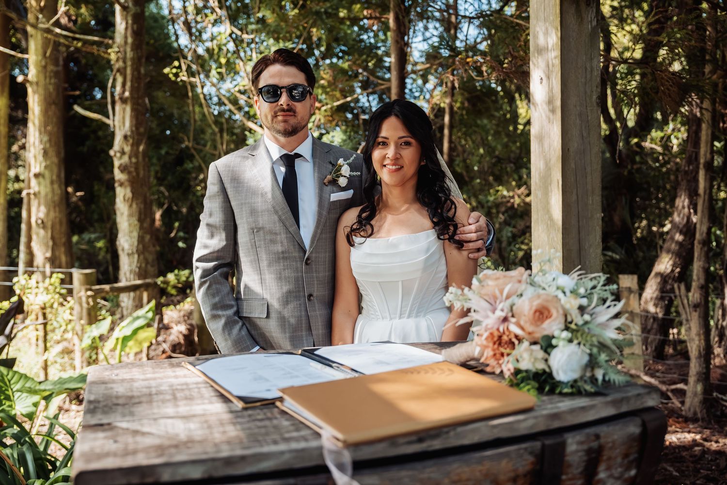 Bride and groom pose together at signing table surrounded by greenery after outdoor woodland ceremony.