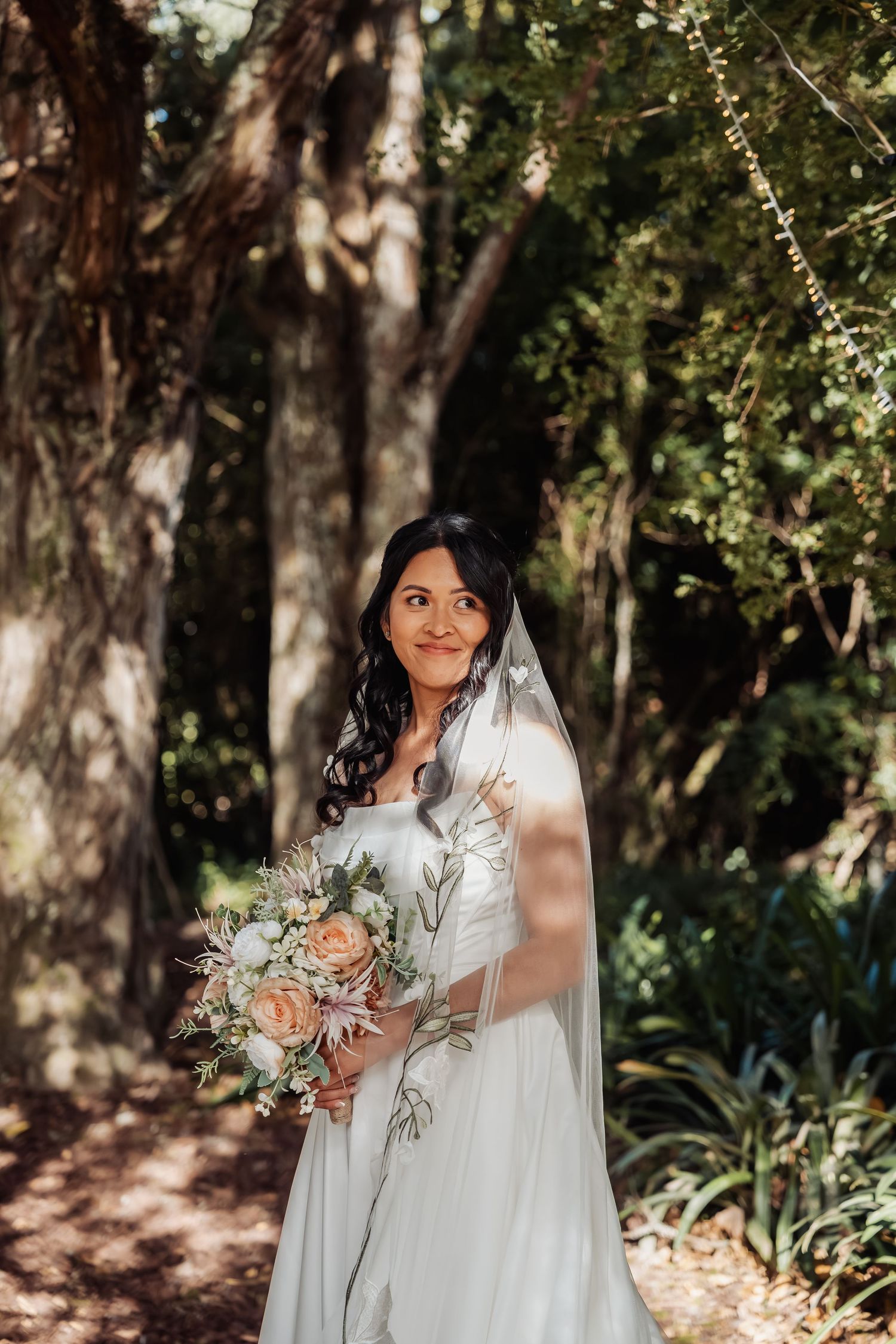 Radiant bride in white gown and veil holds a soft peach and white bouquet in a sunlit forest.