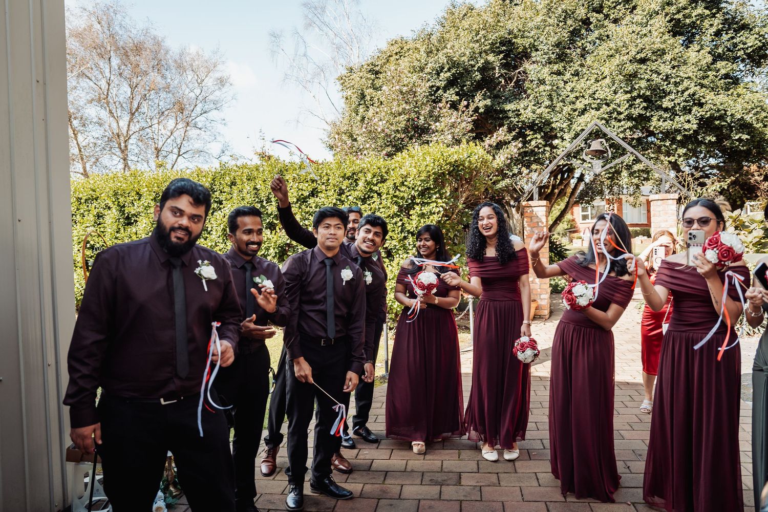 Wedding party in burgundy attire celebrating outdoors, groomsmen in black suits and bridesmaids in maroon dresses.