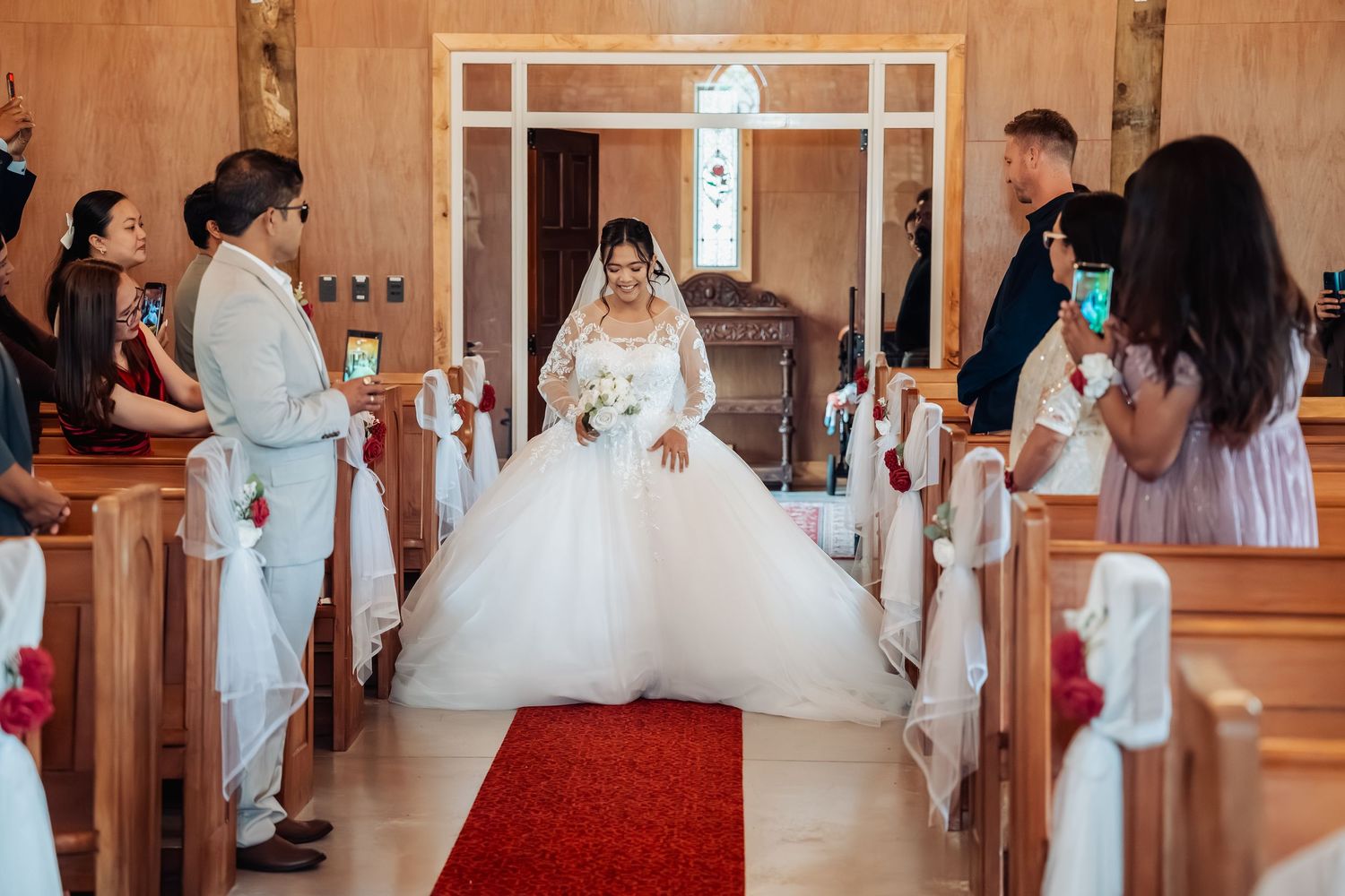 Bride in a stunning white ball gown walks down the red carpet aisle of a traditional church during her wedding ceremony.