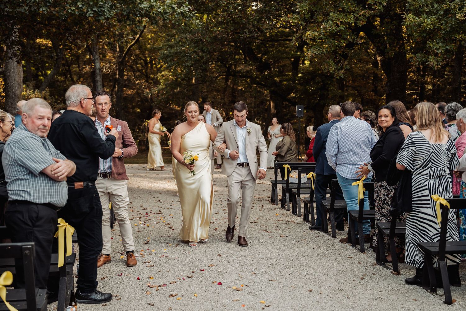 Bride in satin gown and groom in tan suit walk back down the aisle after their outdoor woodland ceremony.