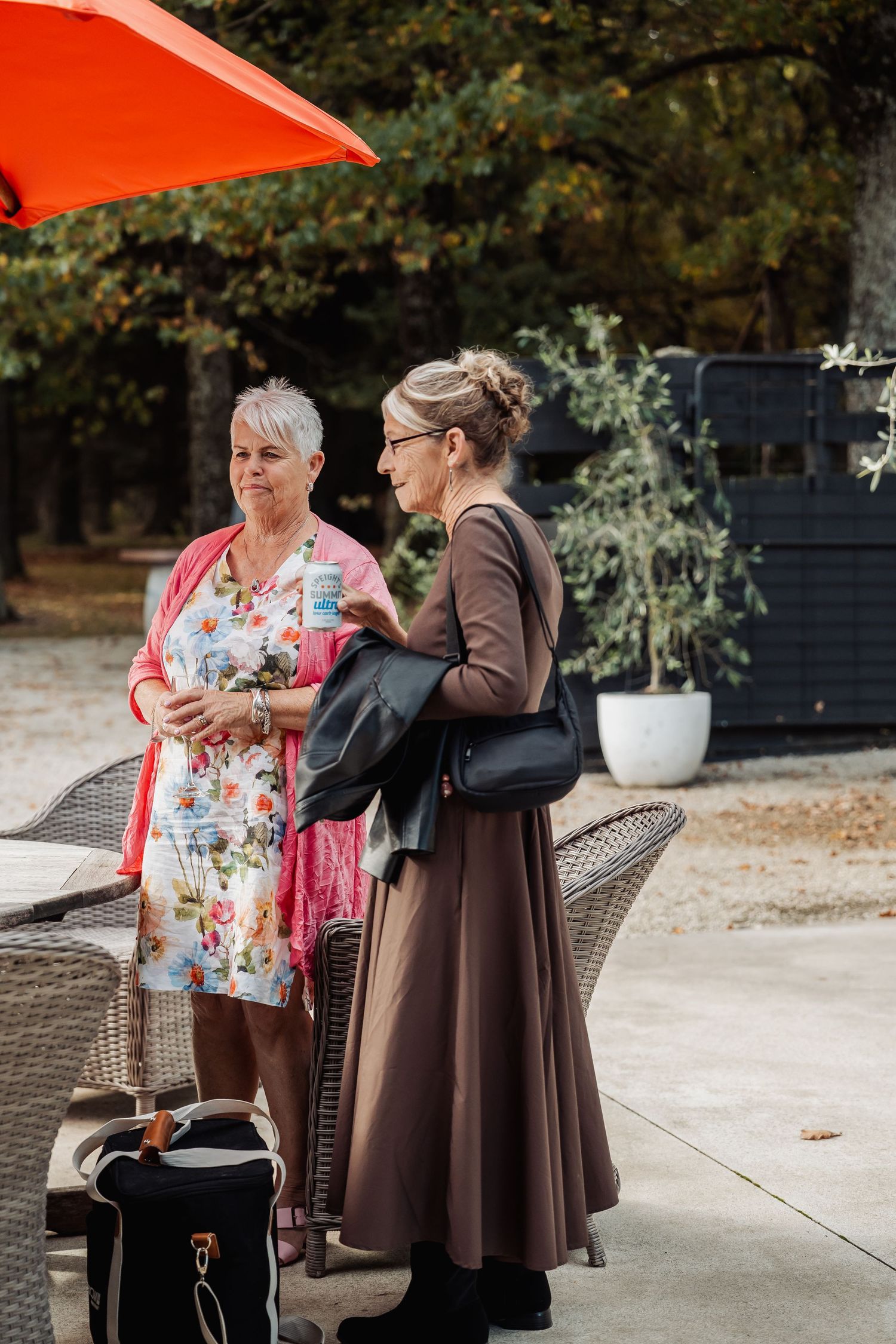 Two elegantly dressed women conversing outdoors under a red umbrella at a wedding venue.