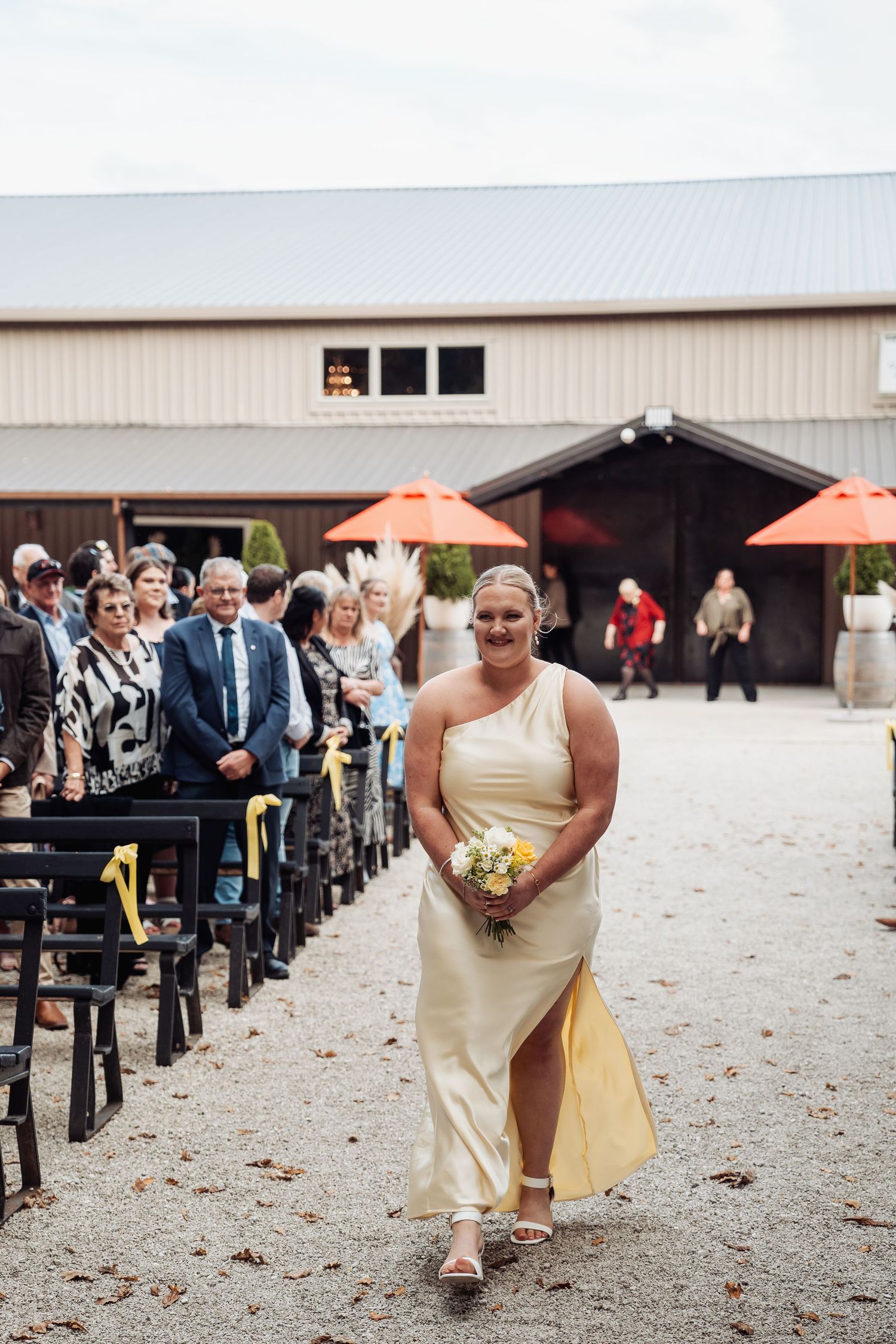 Bridesmaid in yellow dress walks down outdoor aisle holding bouquet before guests.