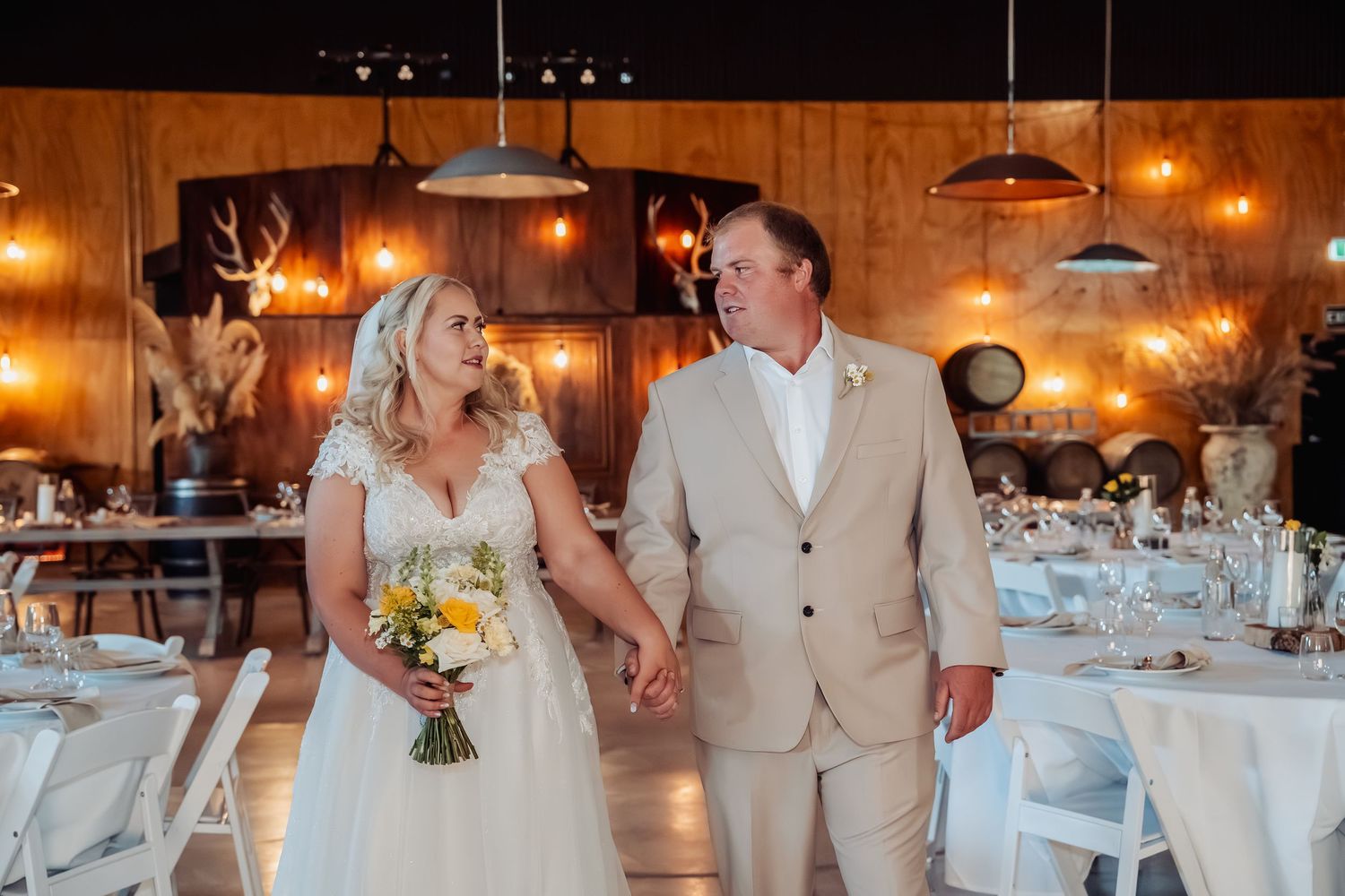 Bride and groom hold hands smiling in rustic barn wedding reception with warm string lights and elegant table settings.