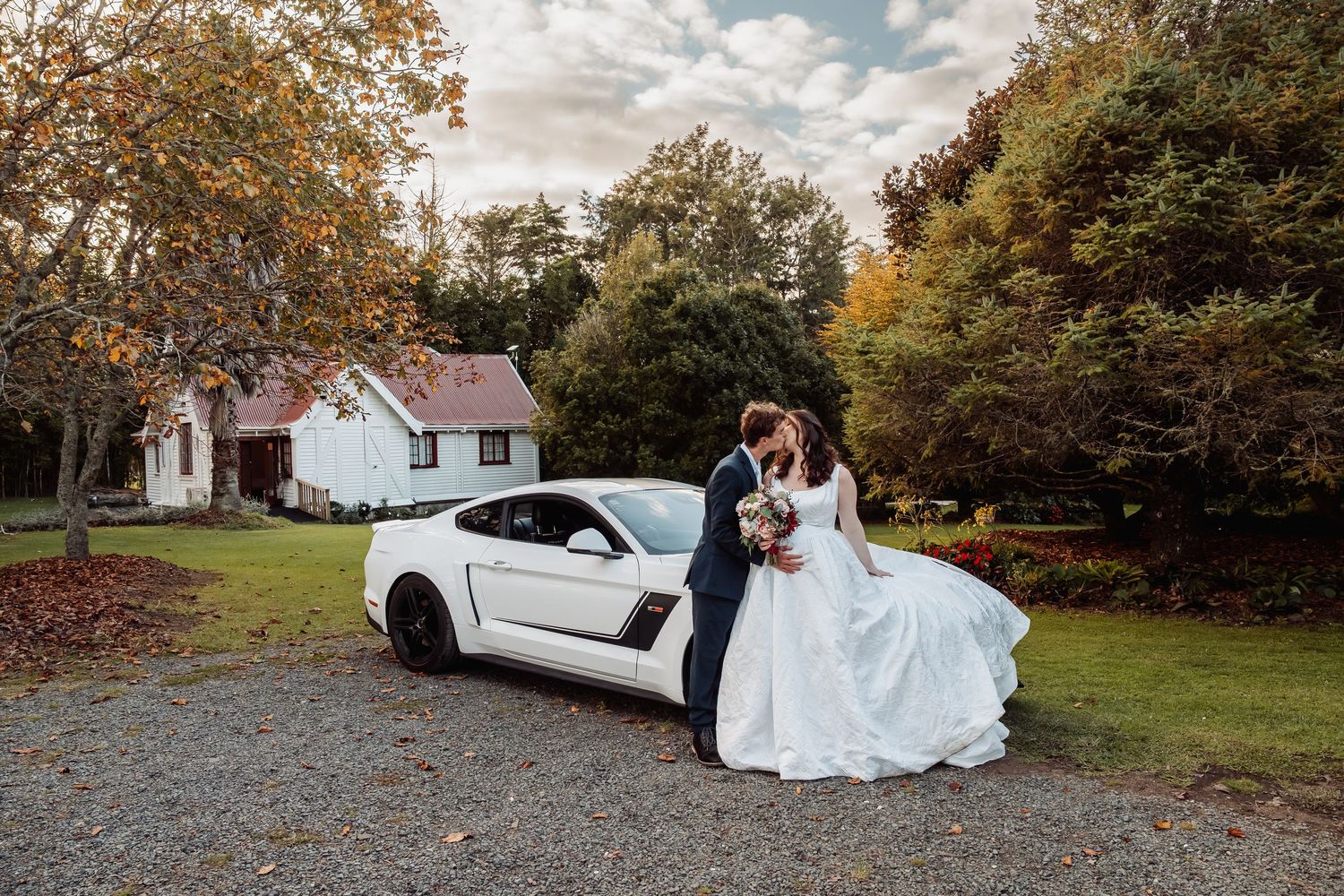 Bride and groom kissing beside a white Ford Mustang on a gravel drive surrounded by autumn foliage.