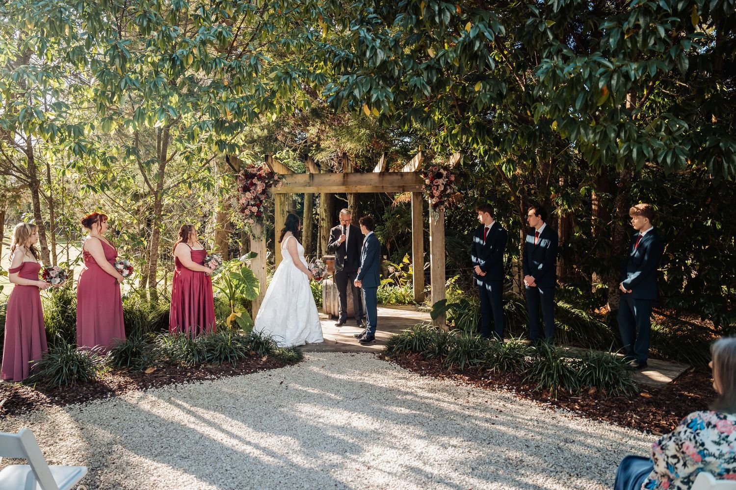 Wedding ceremony in garden with bridesmaids in burgundy dresses and groomsmen in navy suits.