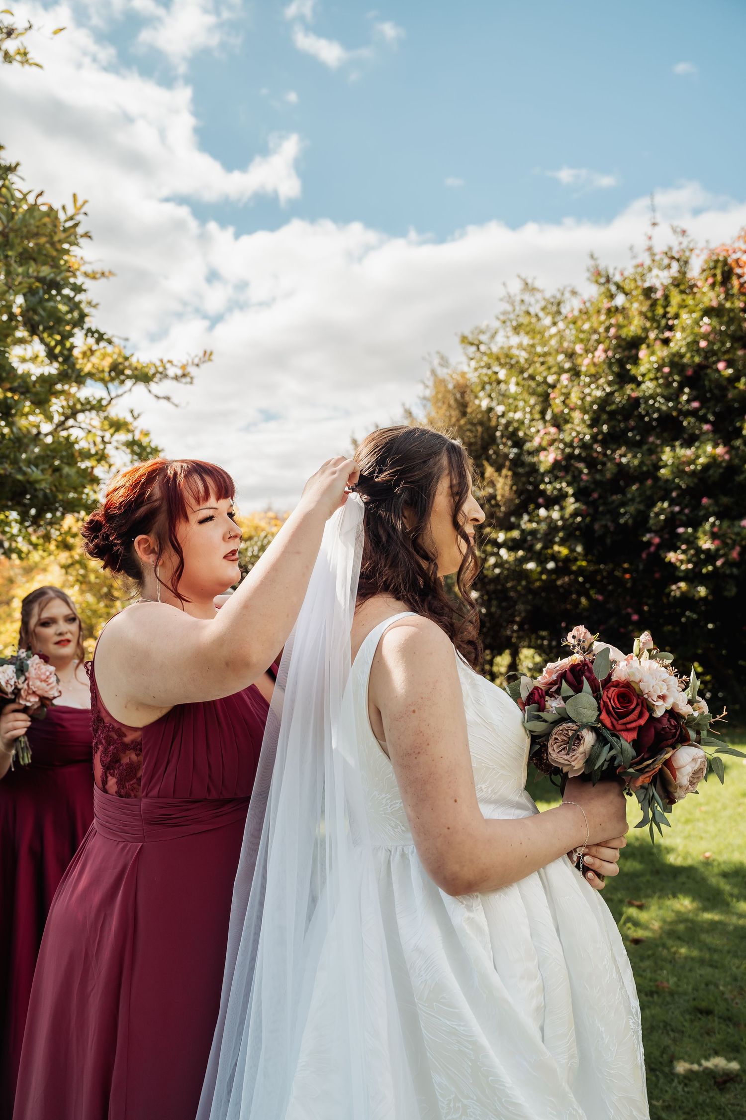 Bridesmaid in burgundy dress adjusting bride's veil outdoors on sunny wedding day.