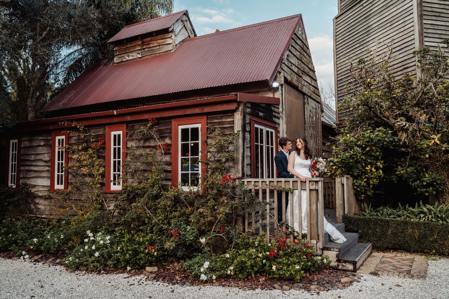 Bride and groom embrace on porch of rustic wooden chapel with red roof.
