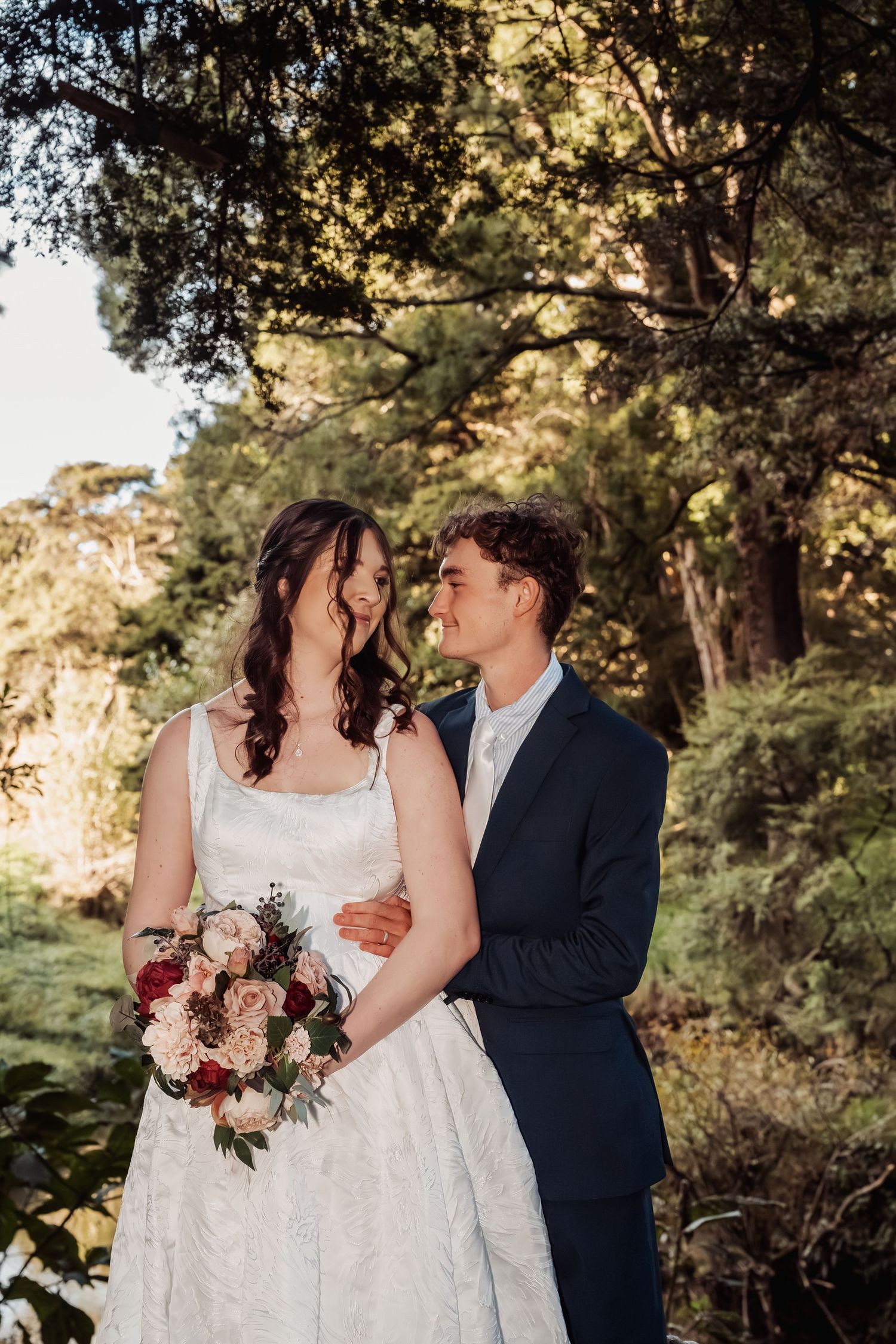 Bride and groom embracing in sunlit forest garden setting.