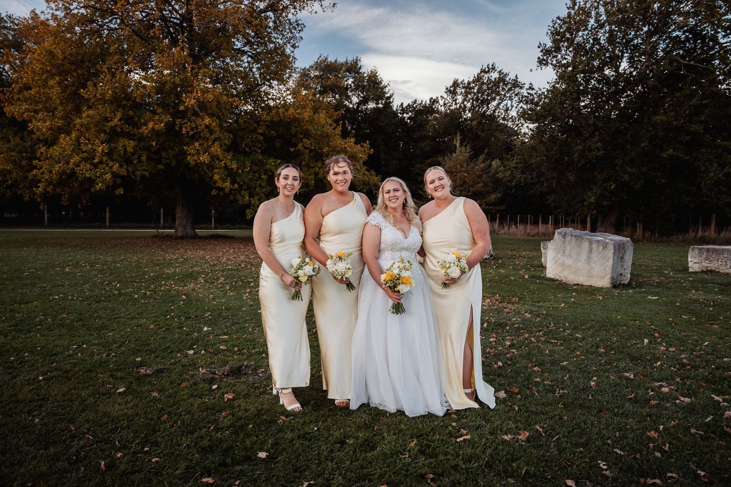 A bride in white stands with three bridesmaids in yellow dresses holding bouquets on a green lawn at sunset.
