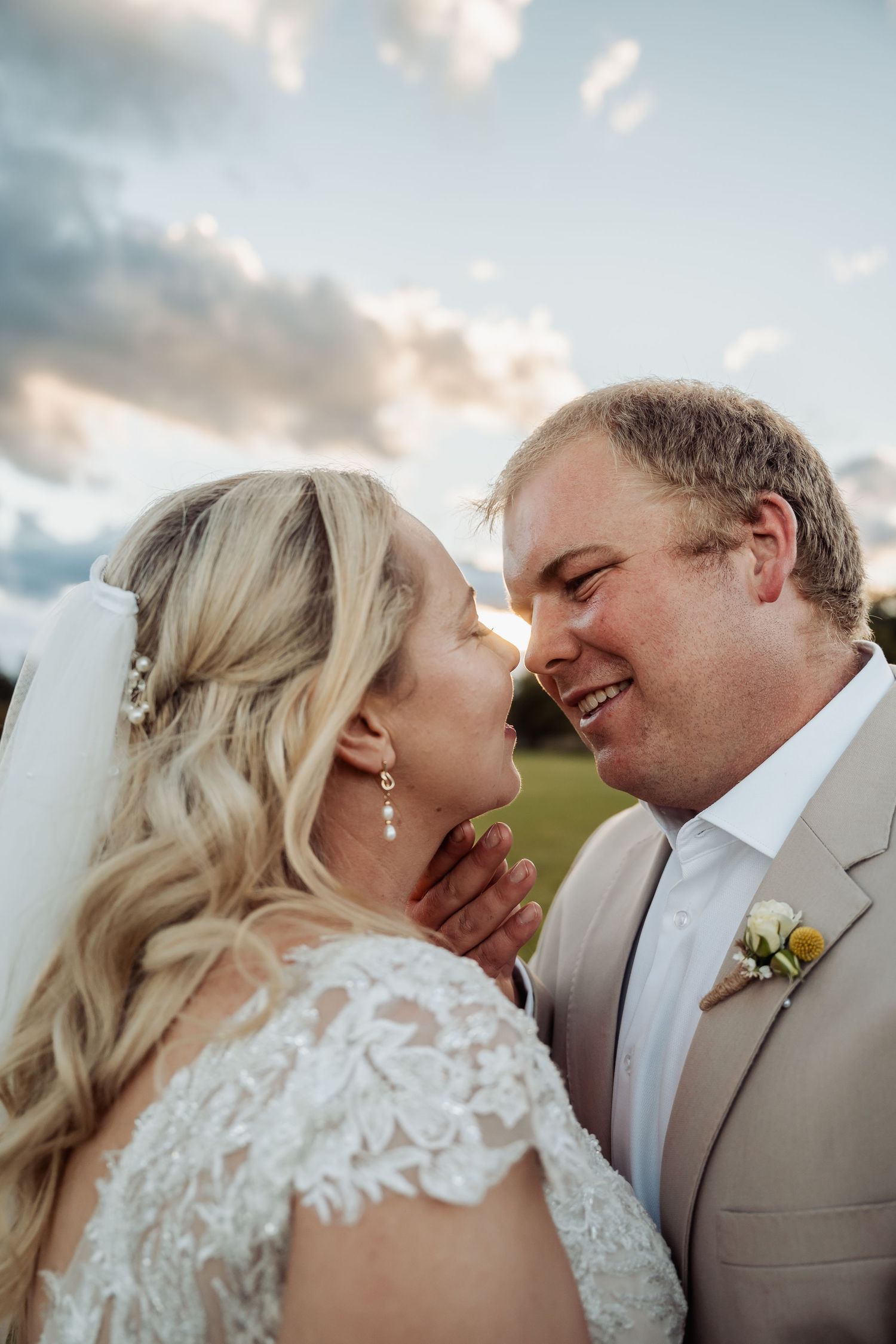 Groom in tan suit tenderly holds bride's face while smiling at her outdoors at sunset.