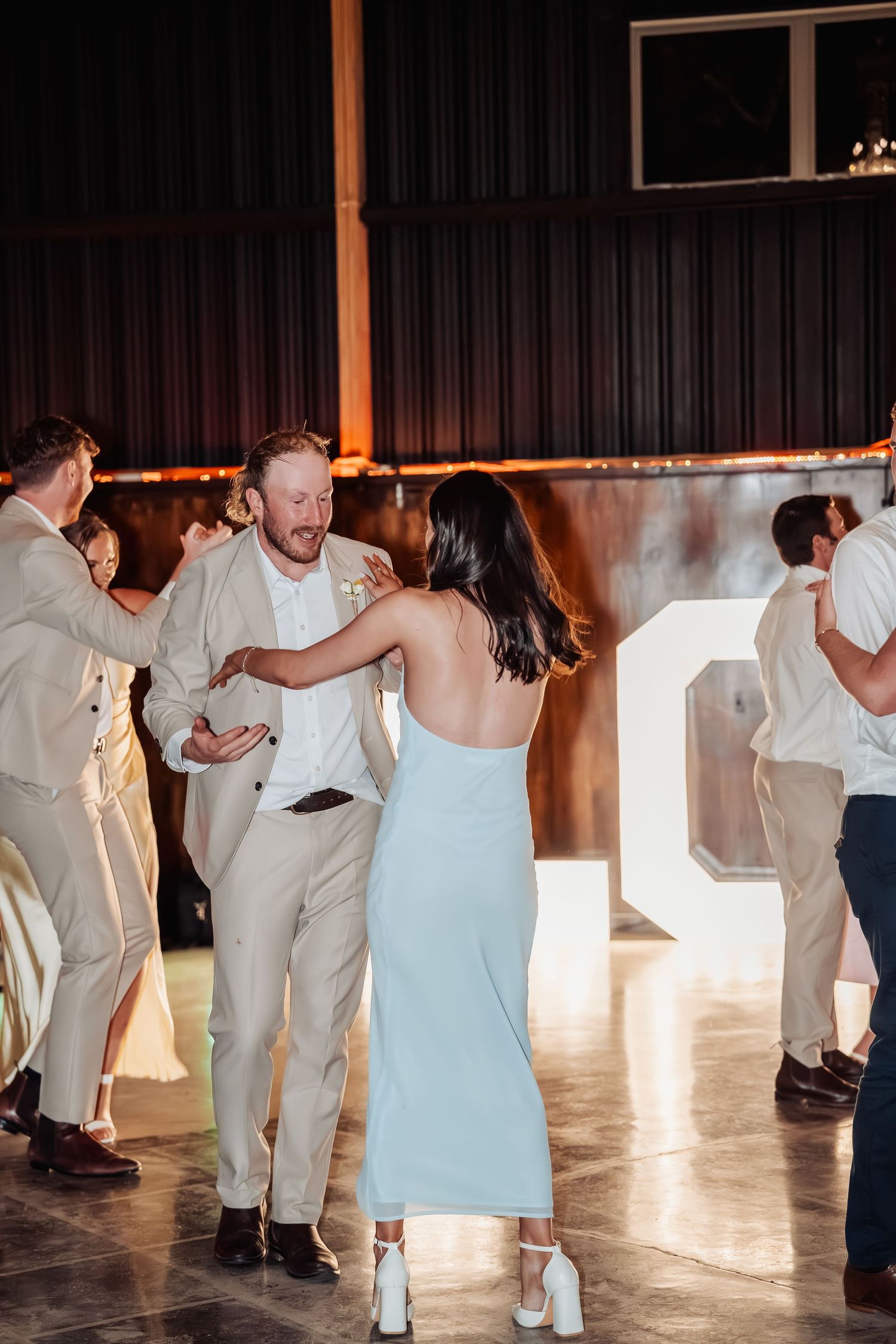 Guests dancing at a wedding reception in a barn venue with string lights and large letter decorations in the background.