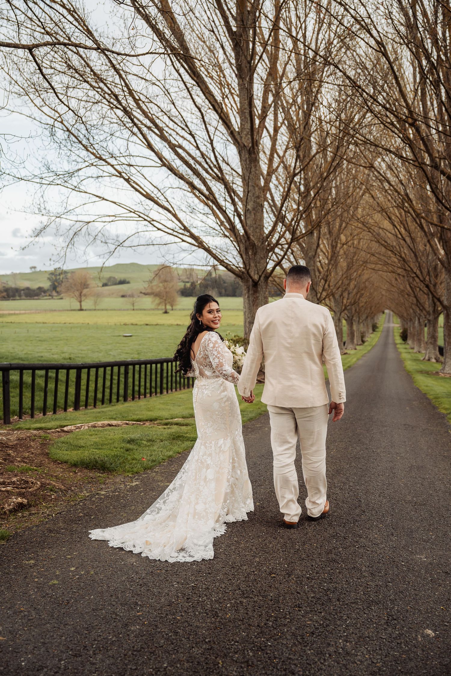 Newlyweds walking down a tree-lined path together in winter landscape.