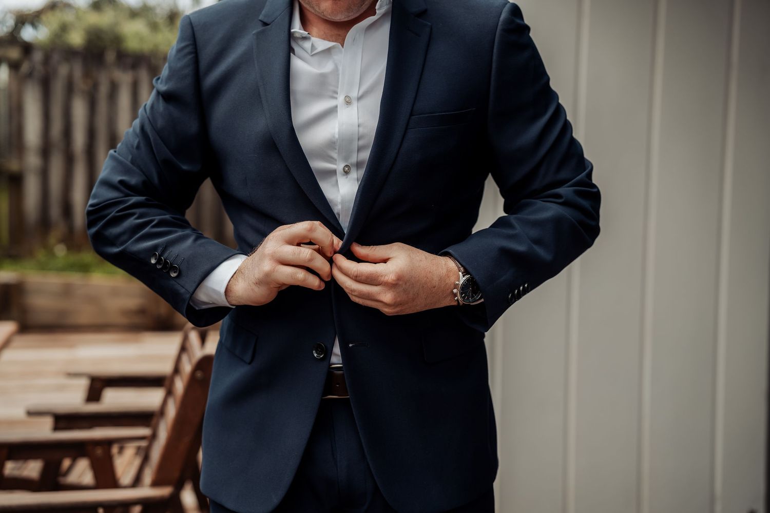 Professional wearing a navy suit jacket adjusts their tie while standing outside near wooden benches.