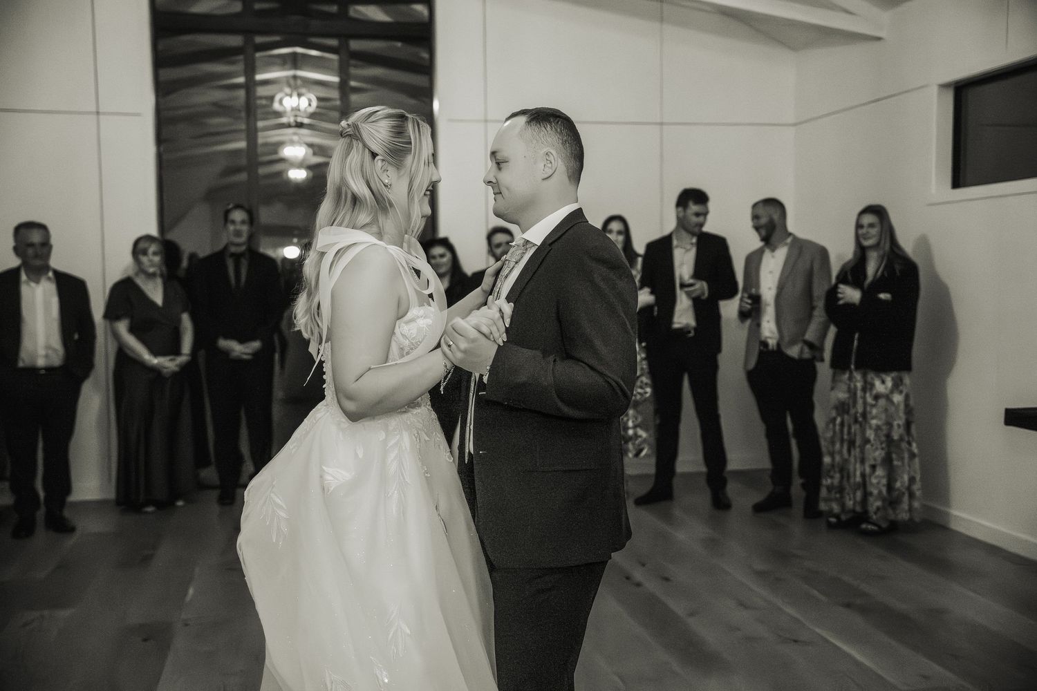 A couple shares their first dance surrounded by guests at an indoor reception venue.