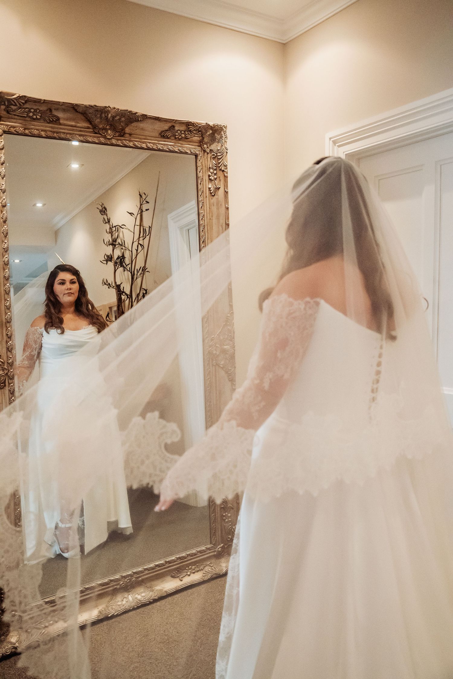 A bridal gown reflection in an ornate vintage mirror shows flowing white dress and veil in a dressing room.
