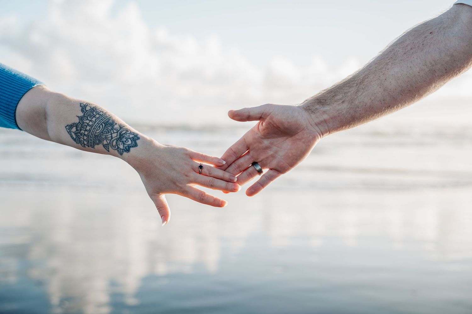 Two hands reaching towards each other against a cloudy sky, one arm adorned with intricate blue tattoo artwork.