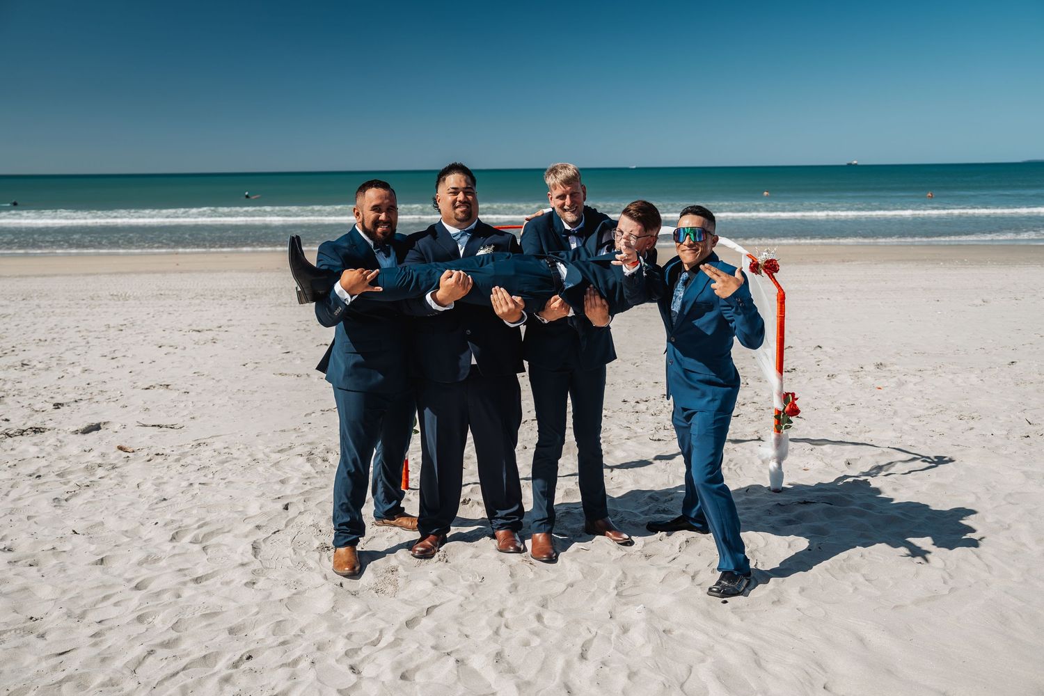 Groomsmen in navy suits pose playfully on a sunny beach.