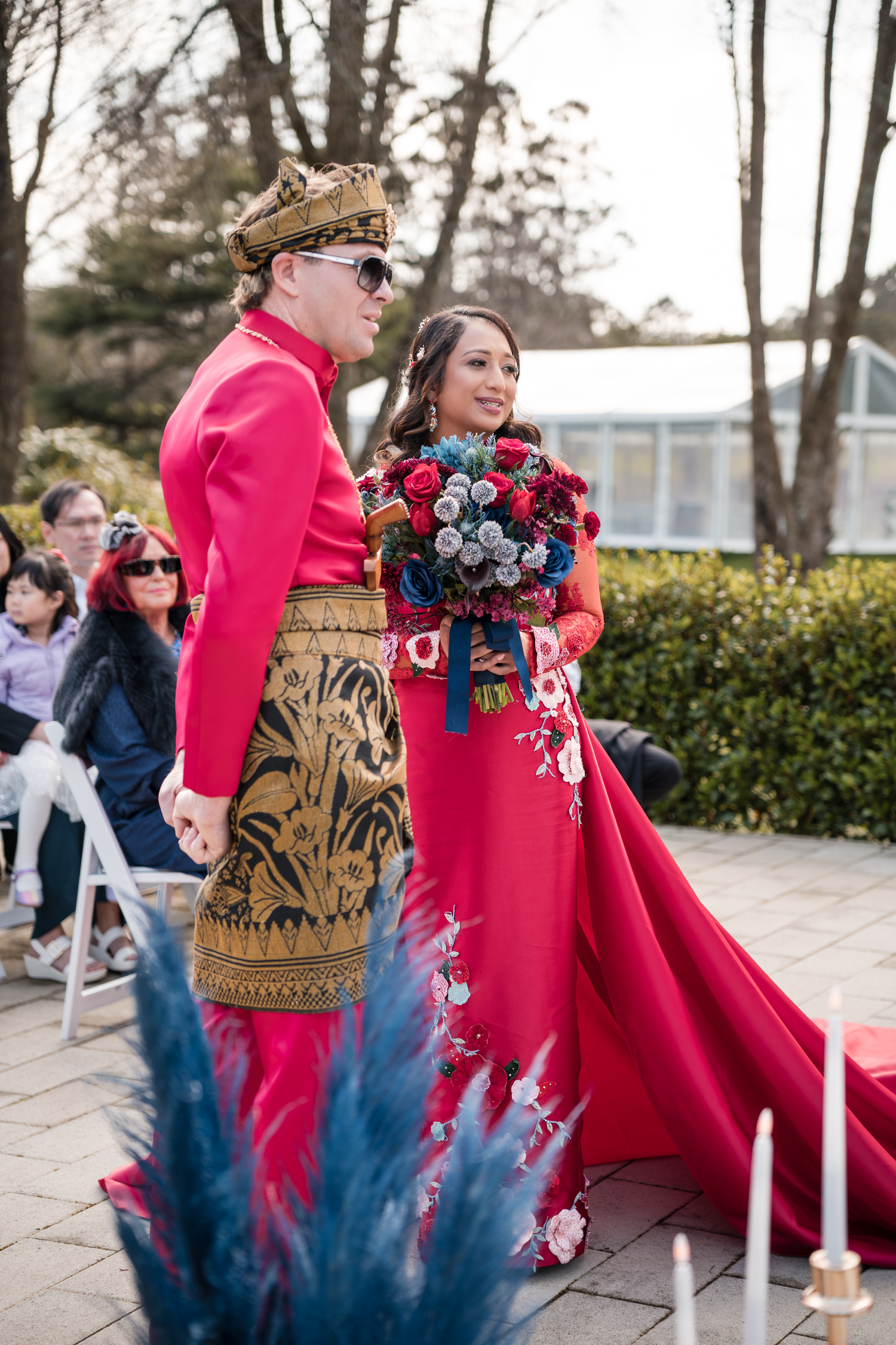 Couple in matching red attire with ornate gold details walking along a decorated outdoor ceremony space.
