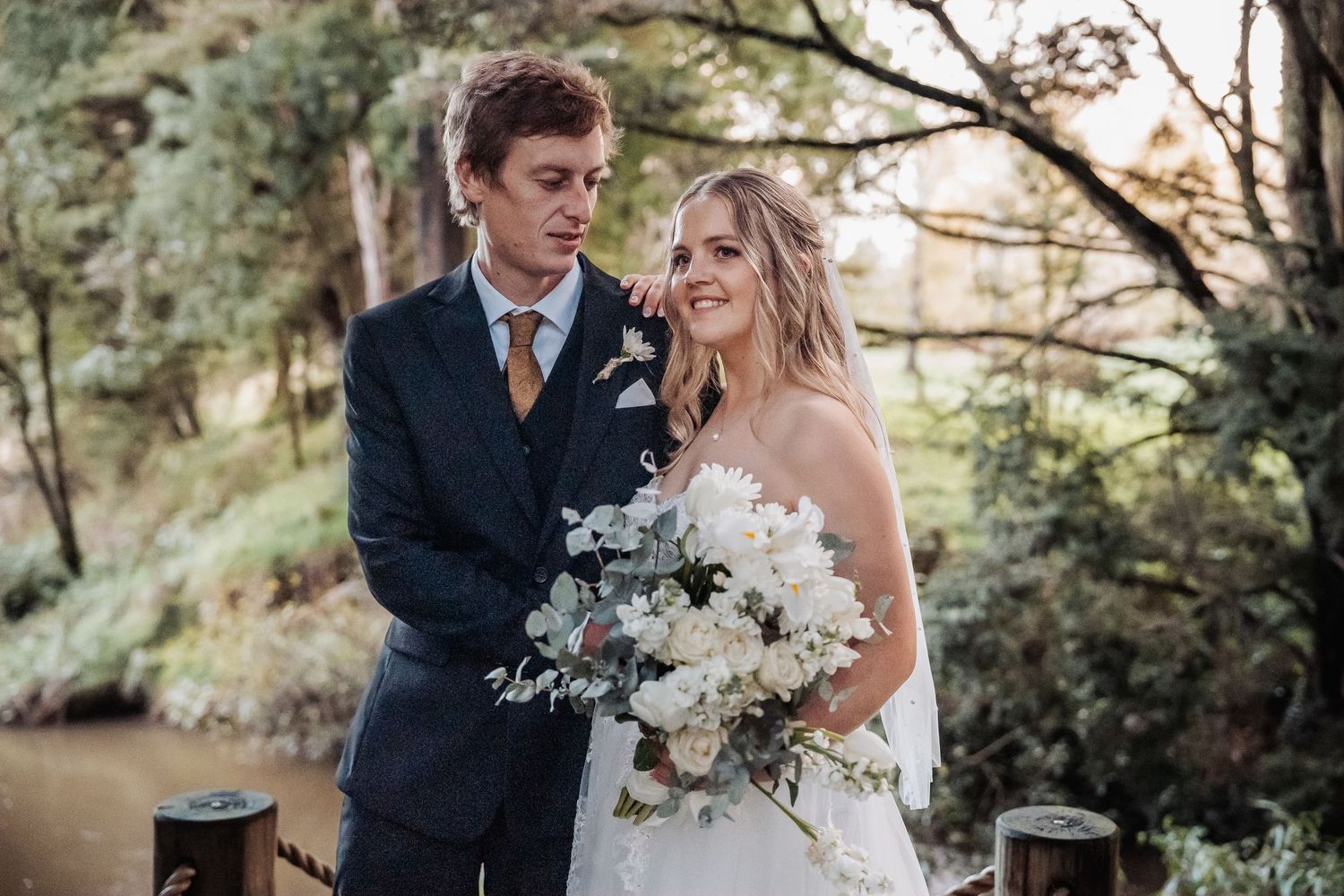 Happy newlyweds in formal attire pose together with elegant white bouquet in outdoor garden setting.