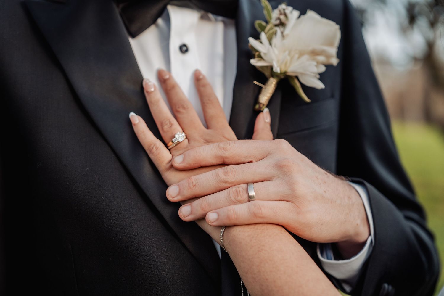 Close-up of wedding rings and hands with a white boutonniere on a black tuxedo lapel.