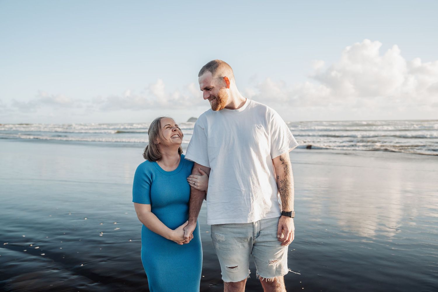 Two people share a tender moment while standing on a beach at sunset with calm ocean waters behind them.