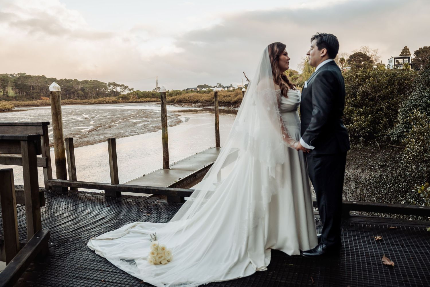A romantic wedding couple stands on a wooden dock overlooking the water at sunset, with the bride's long train flowing behind.