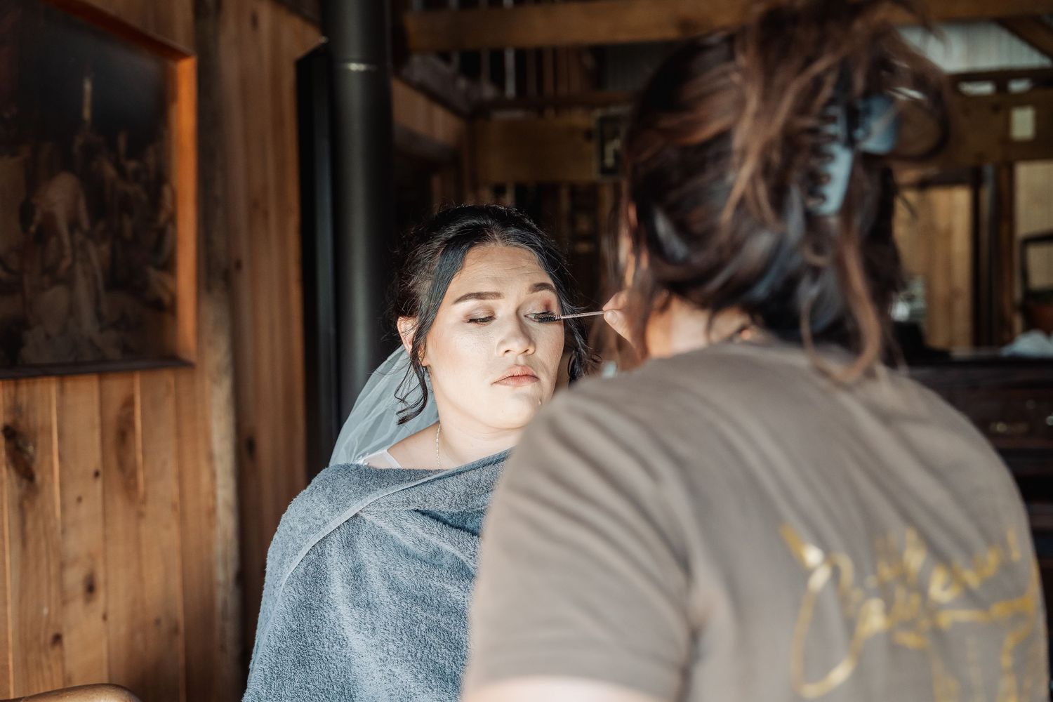 A makeup artist applies cosmetics to a person wearing a white veil in a rustic wood-paneled room.