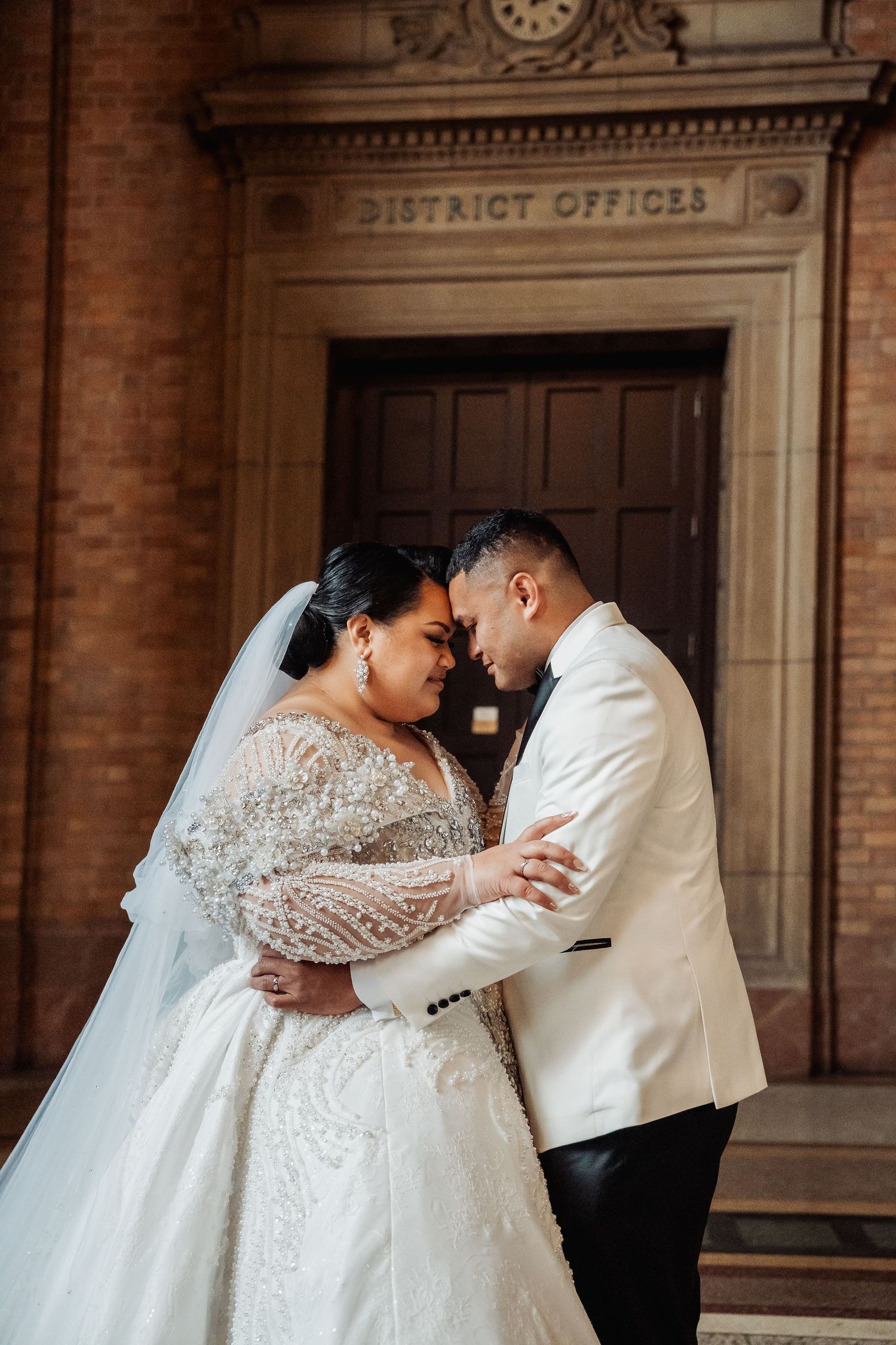 Intimate moment between newlyweds under 'District Offices' sign at historic brick building.
