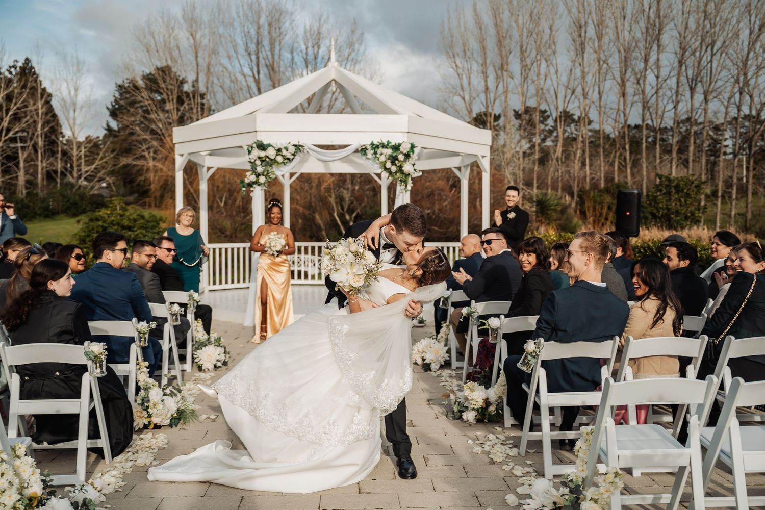 An elegant outdoor wedding ceremony setup with white chairs and a gazebo surrounded by bare winter trees.