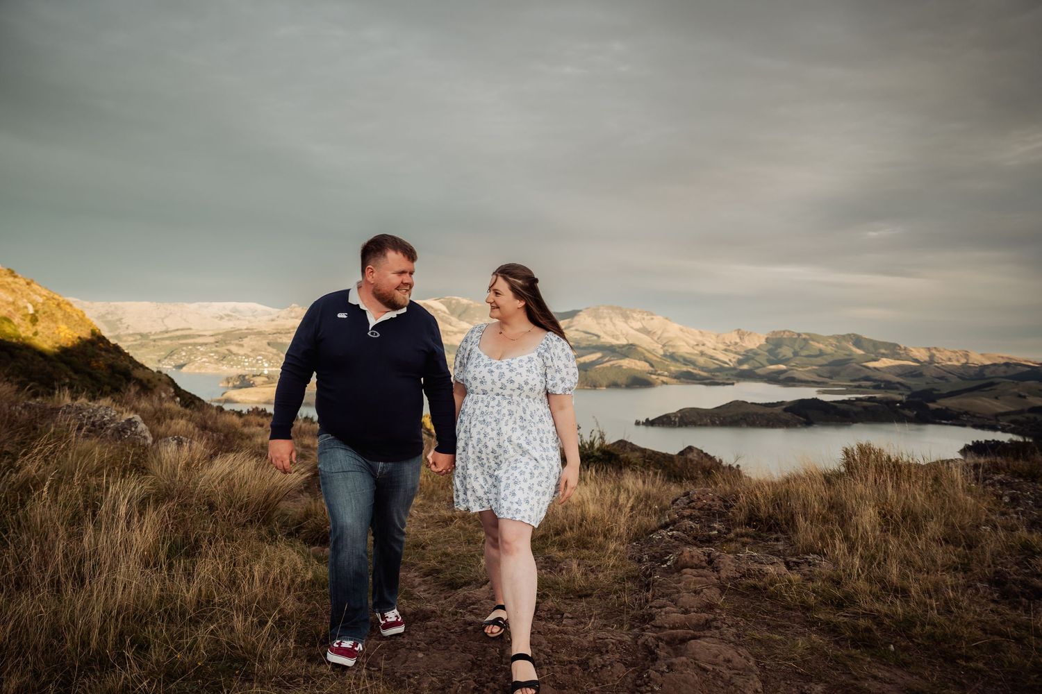 Happy couple walking hand in hand on a scenic hillside with mountains and lake views behind them.