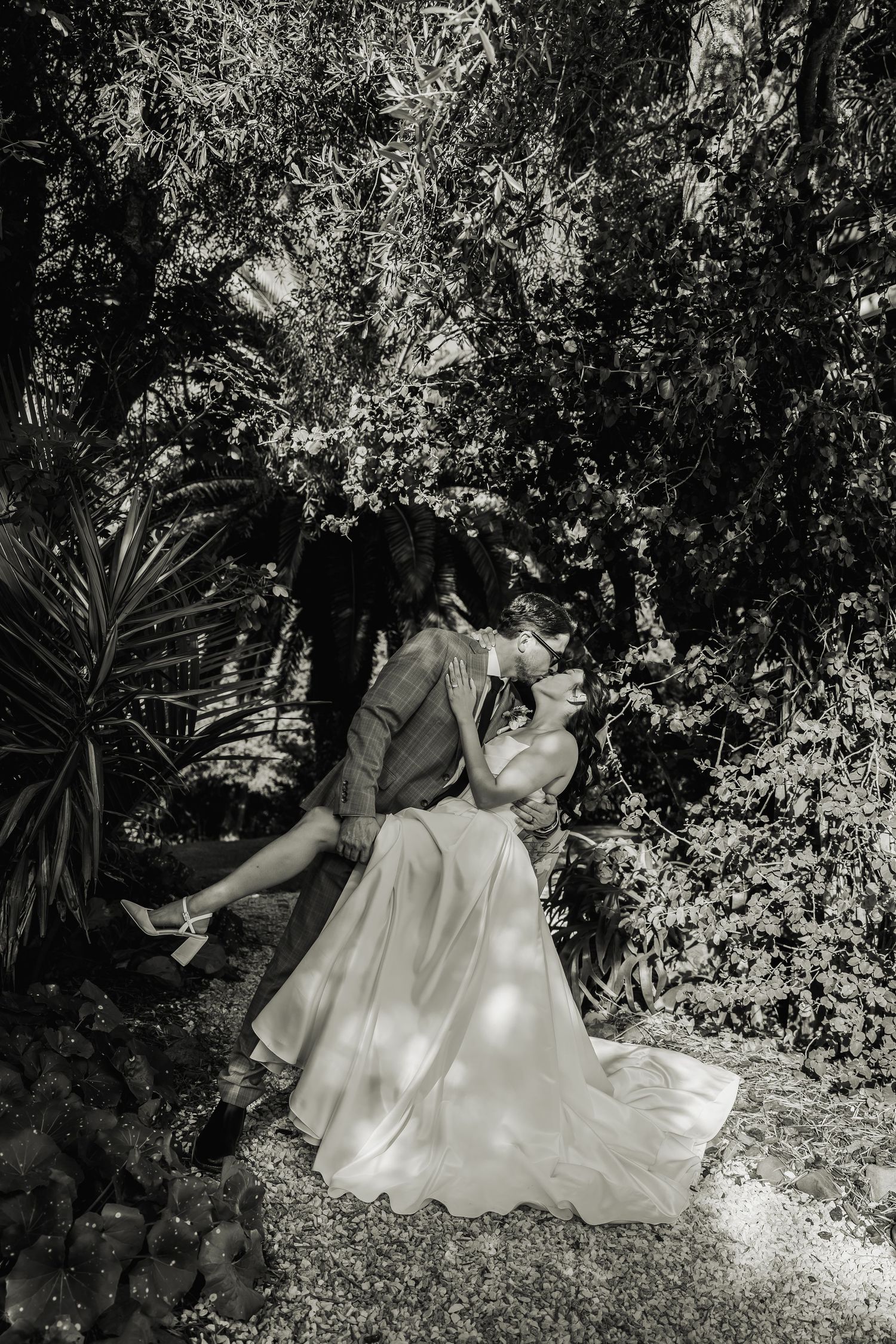 Black and white photo of groom dipping bride in romantic kiss surrounded by lush garden greenery.