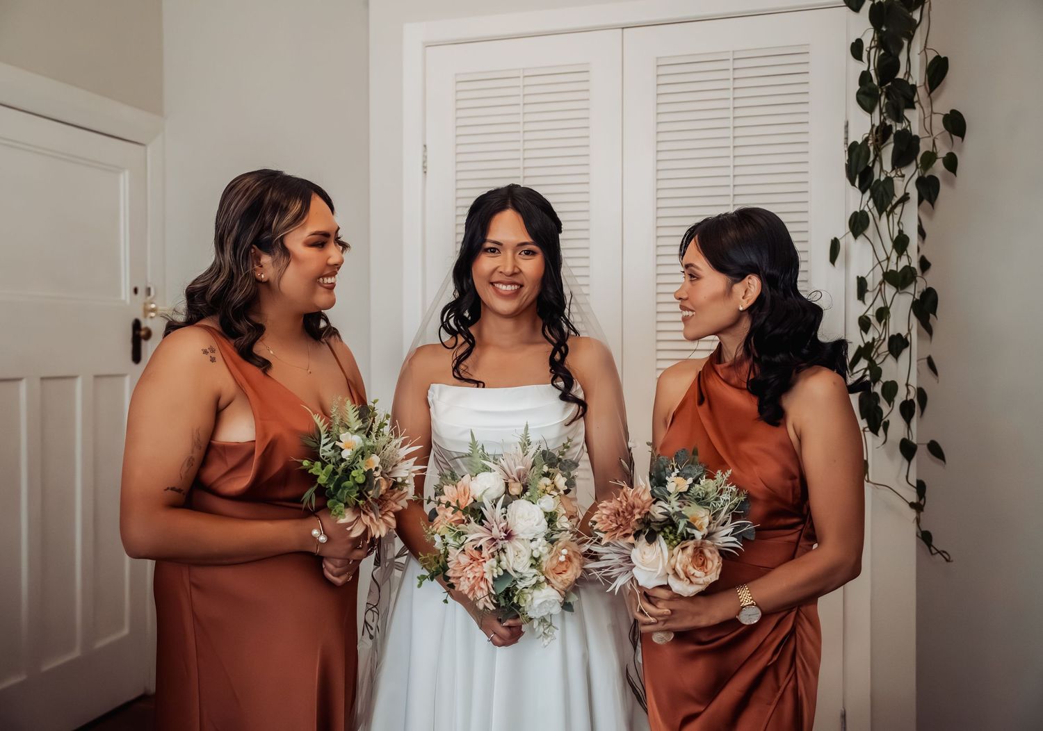 Bride in white gown smiles with two bridesmaids in rust satin dresses holding bouquets indoors.